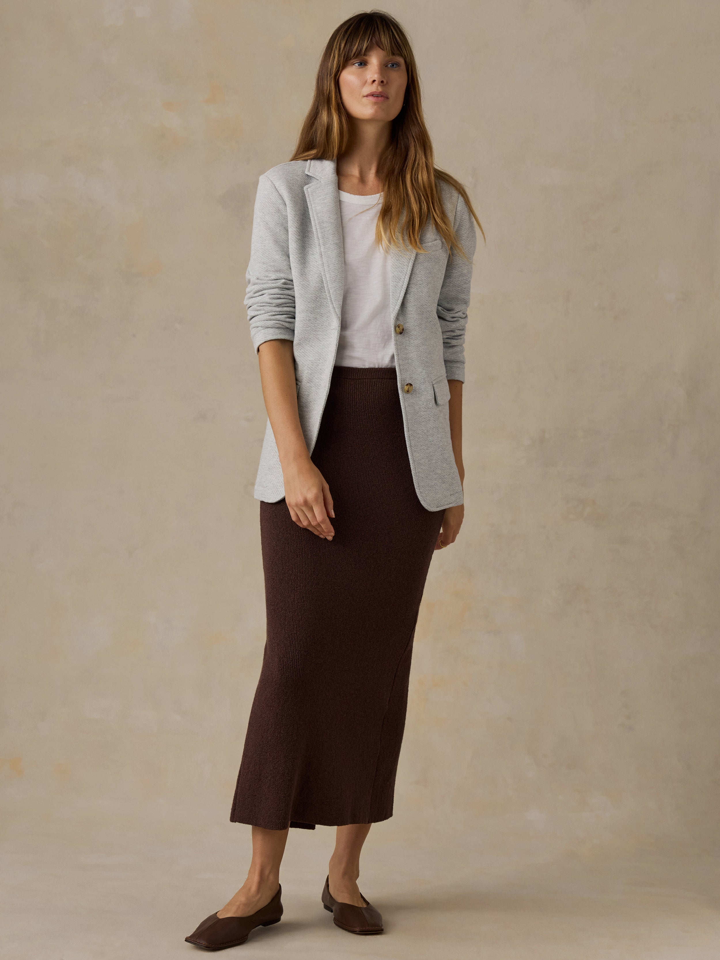 Woman wearing a grey blazer over a white t-shirt and brown skirt against a beige background
