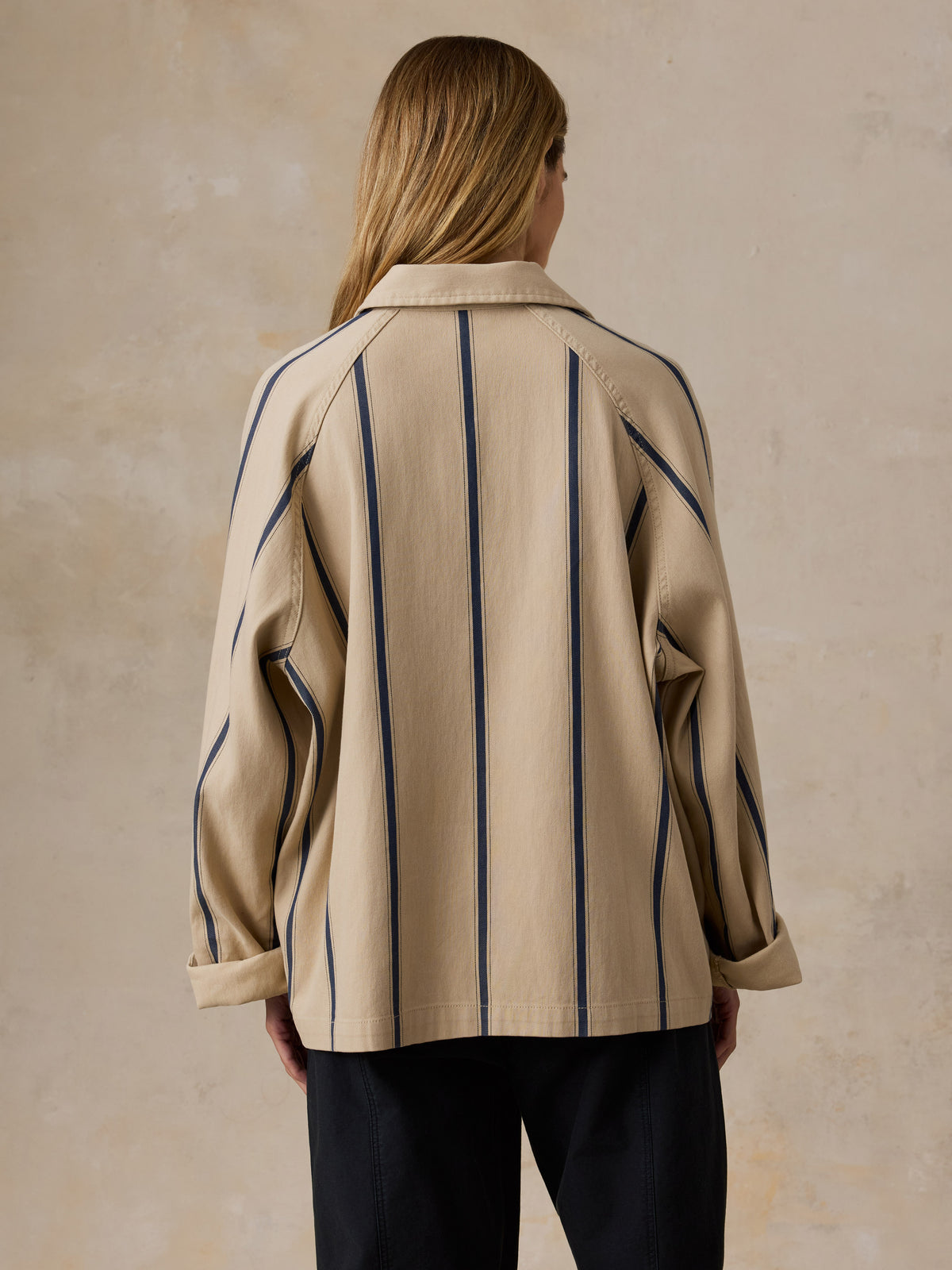 A woman shown from the back wearing a beige button-up shirt with vertical navy blue stripes and dark navy pants, standing against a textured beige wall., image 3 of 7