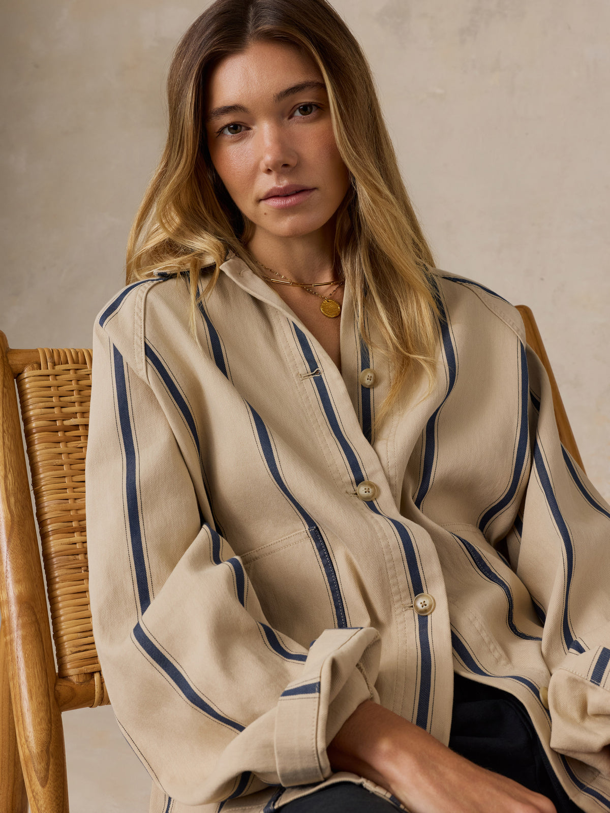 A woman wearing a beige button-up shirt with vertical navy blue stripes, seated on a wooden wicker chair against a textured beige wall., image 5 of 7