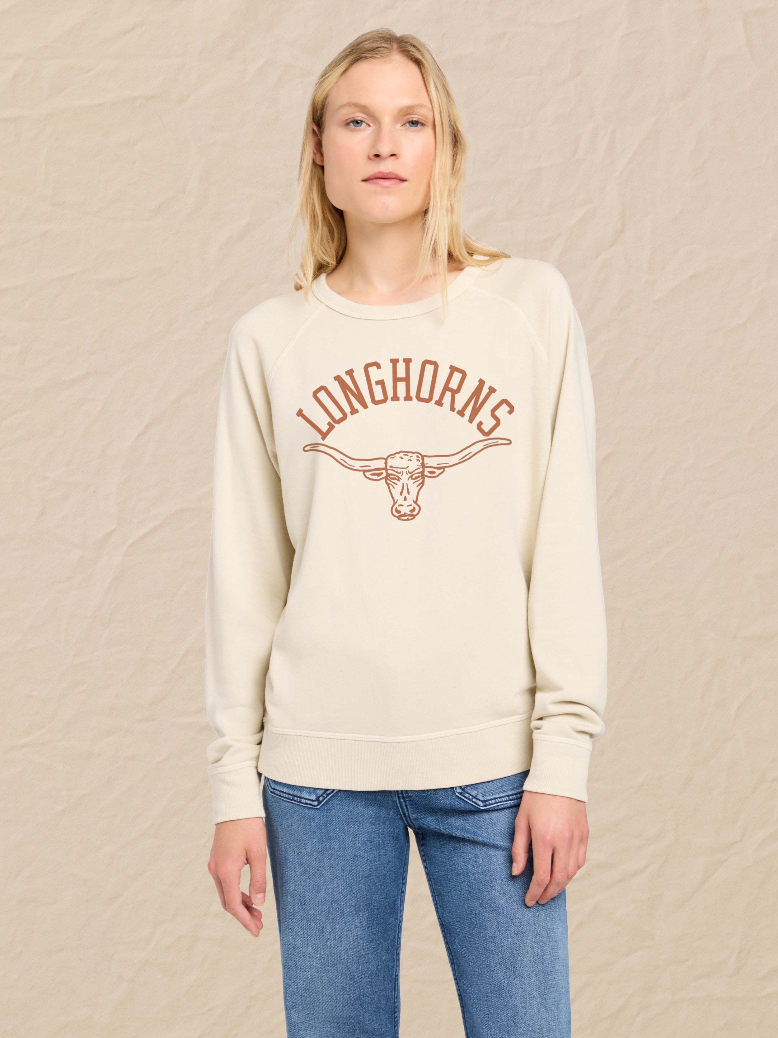 Woman is wearing an ivory crewneck with UT Austin graphic on the center front against a beige background