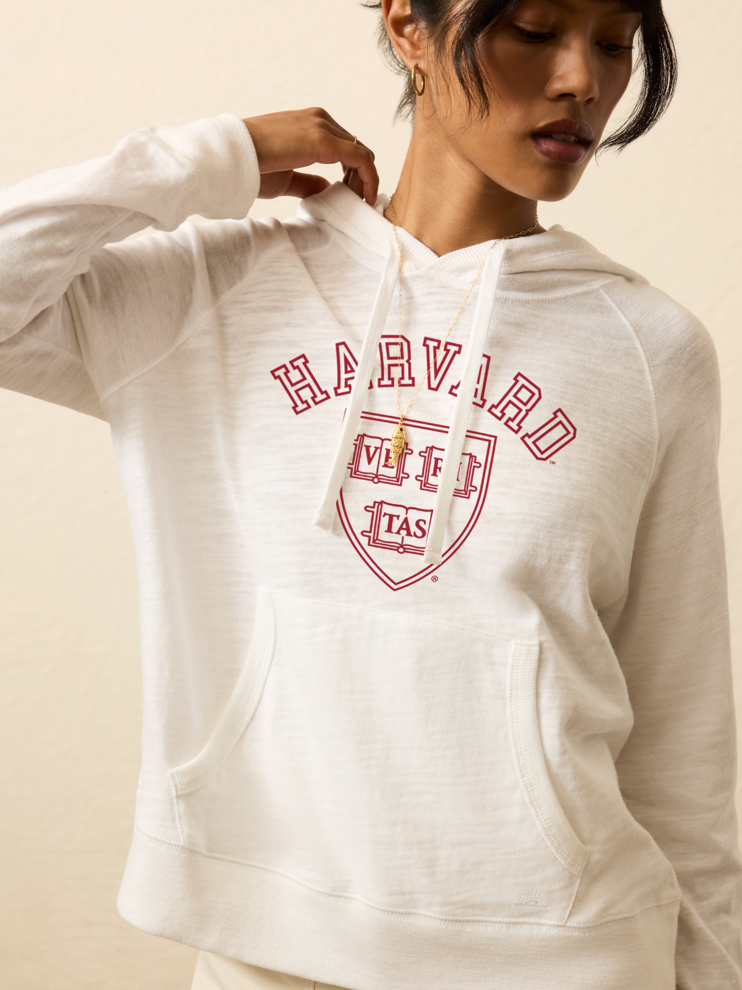Woman is wearing a white hoodie with Harvard graphic on the center front against a beige background