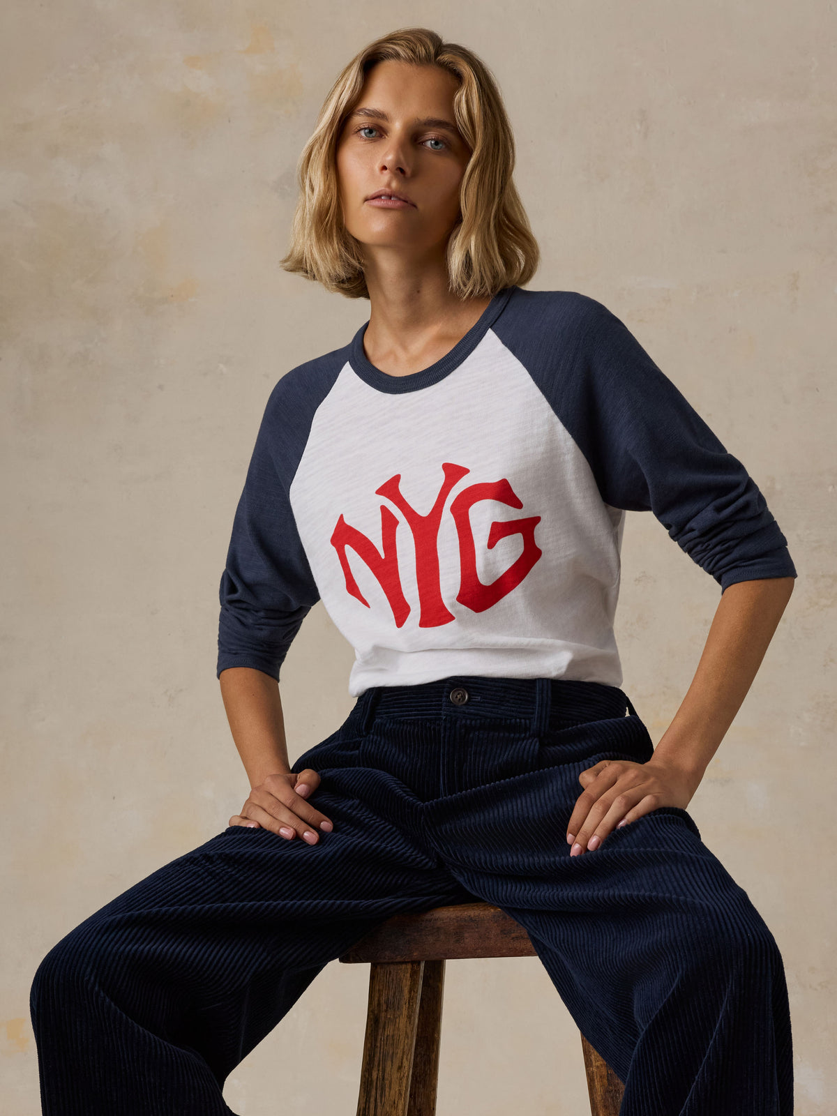 A woman sitting on a stool wearing a white and blue NY Giants baseball tee with navy pants, image 2 of 5