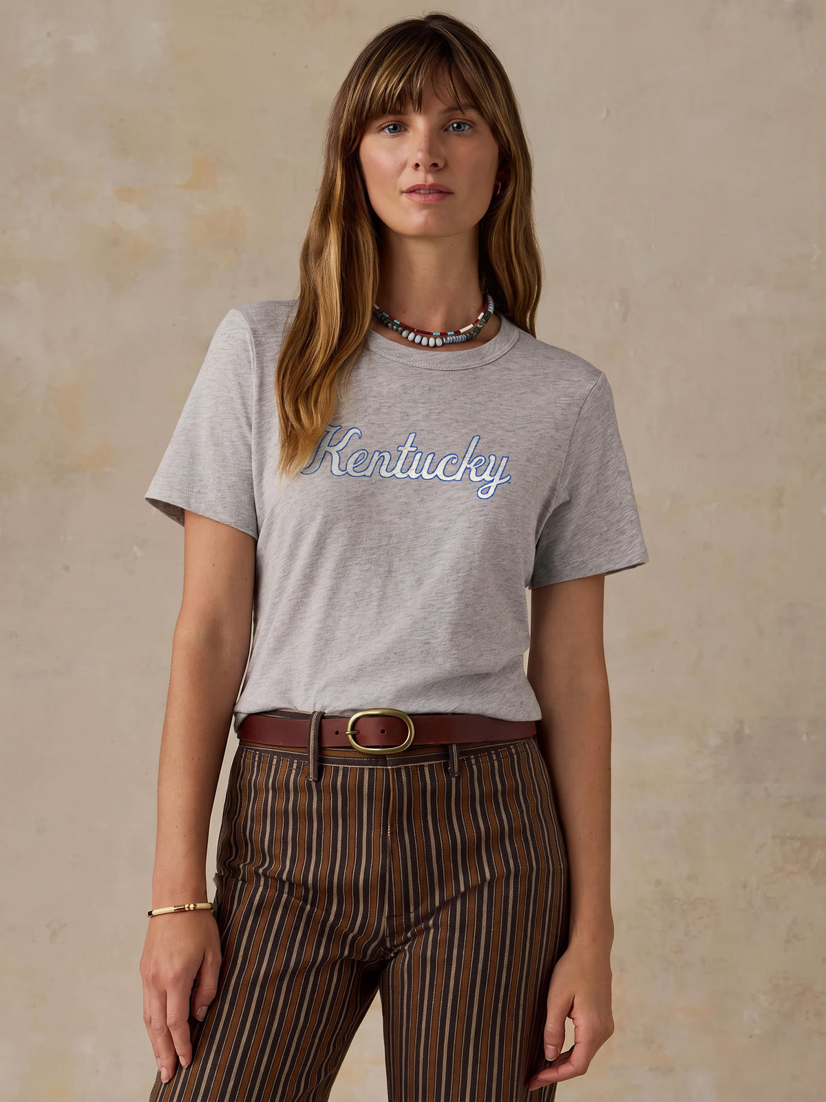 A woman wearing a grey University of Kentucky tee and brown striped pants, image 1 of 1