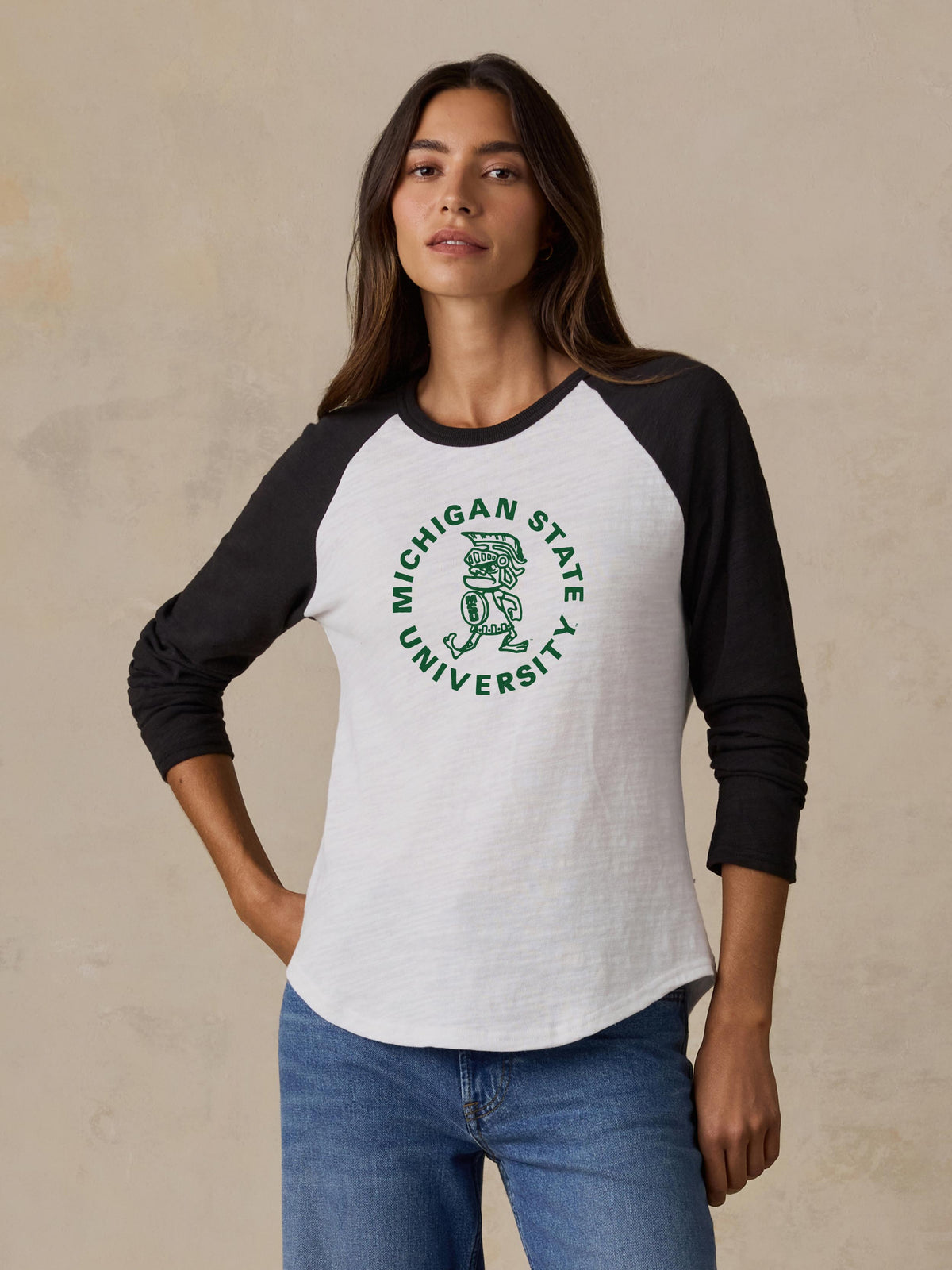 A woman is wearing a black and white Michigan State baseball tee and blue jeans, image 1 of 2
