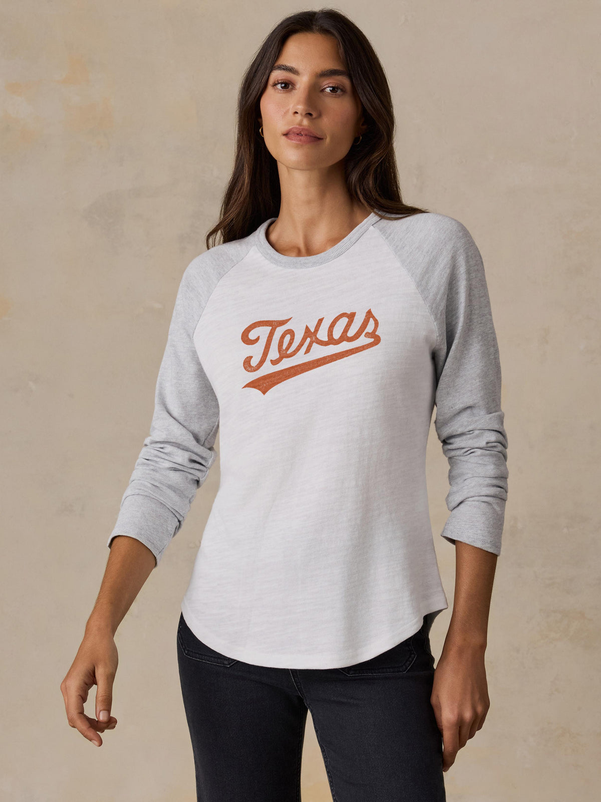 A woman wearing a grey and white UT Austin baseball tee and black jeans, image 1 of 2