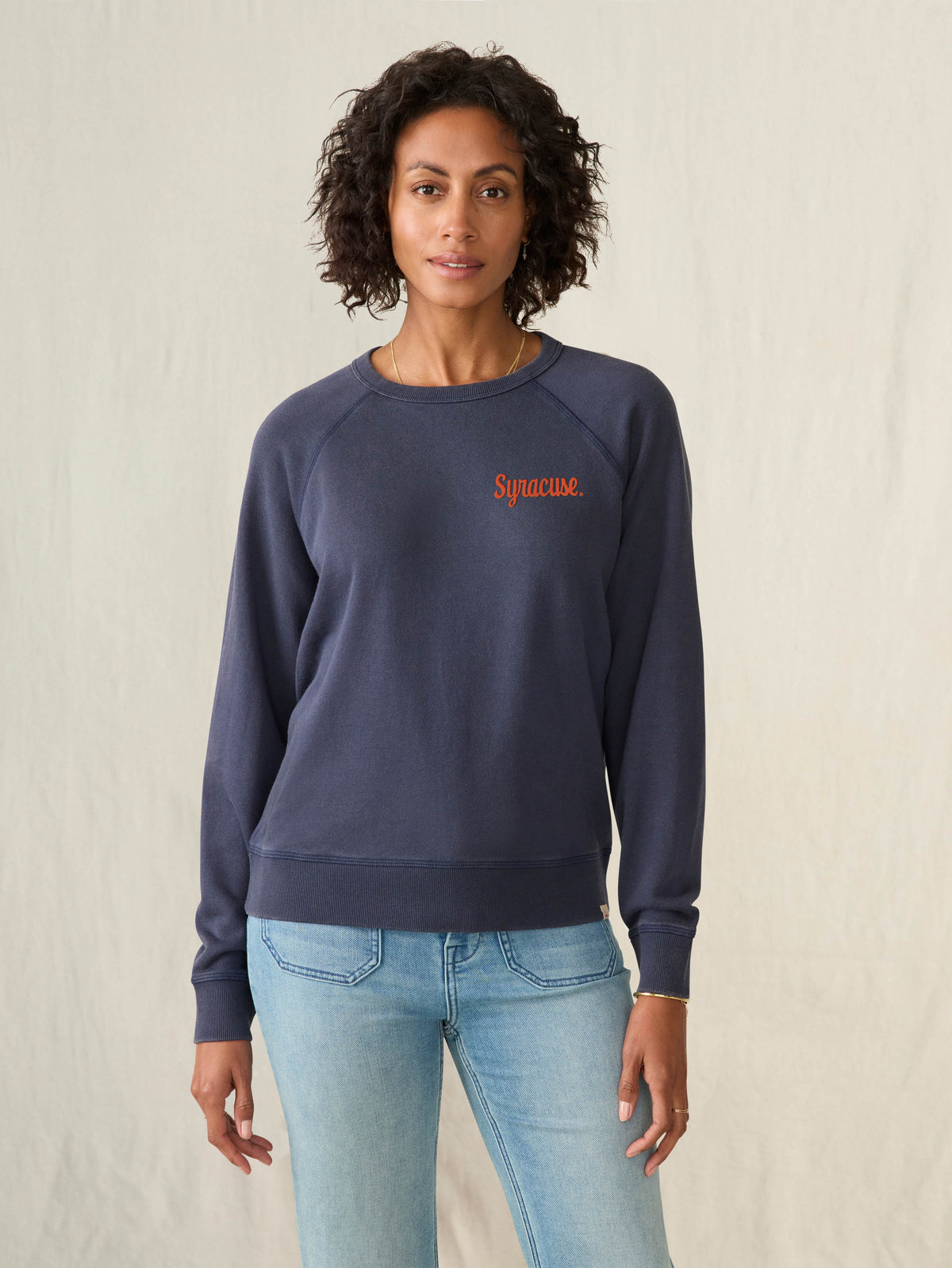 Woman wearing a navy sweatshirt with a Syracuse University graphic on the left chest on a beige background, image 1 of 2