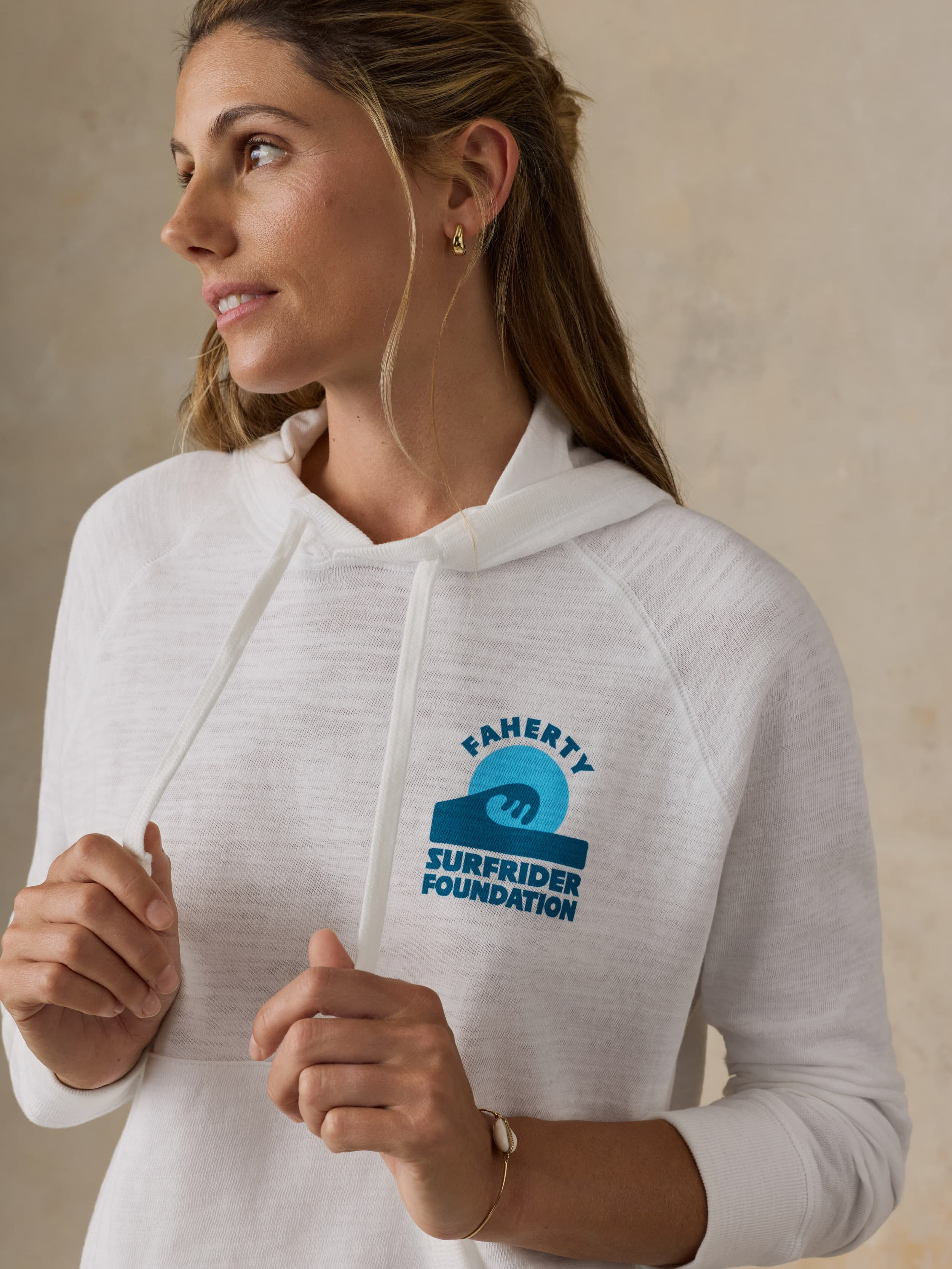 Woman wearing a white Faherty x Surfrider Foundation hoodie against a beige background