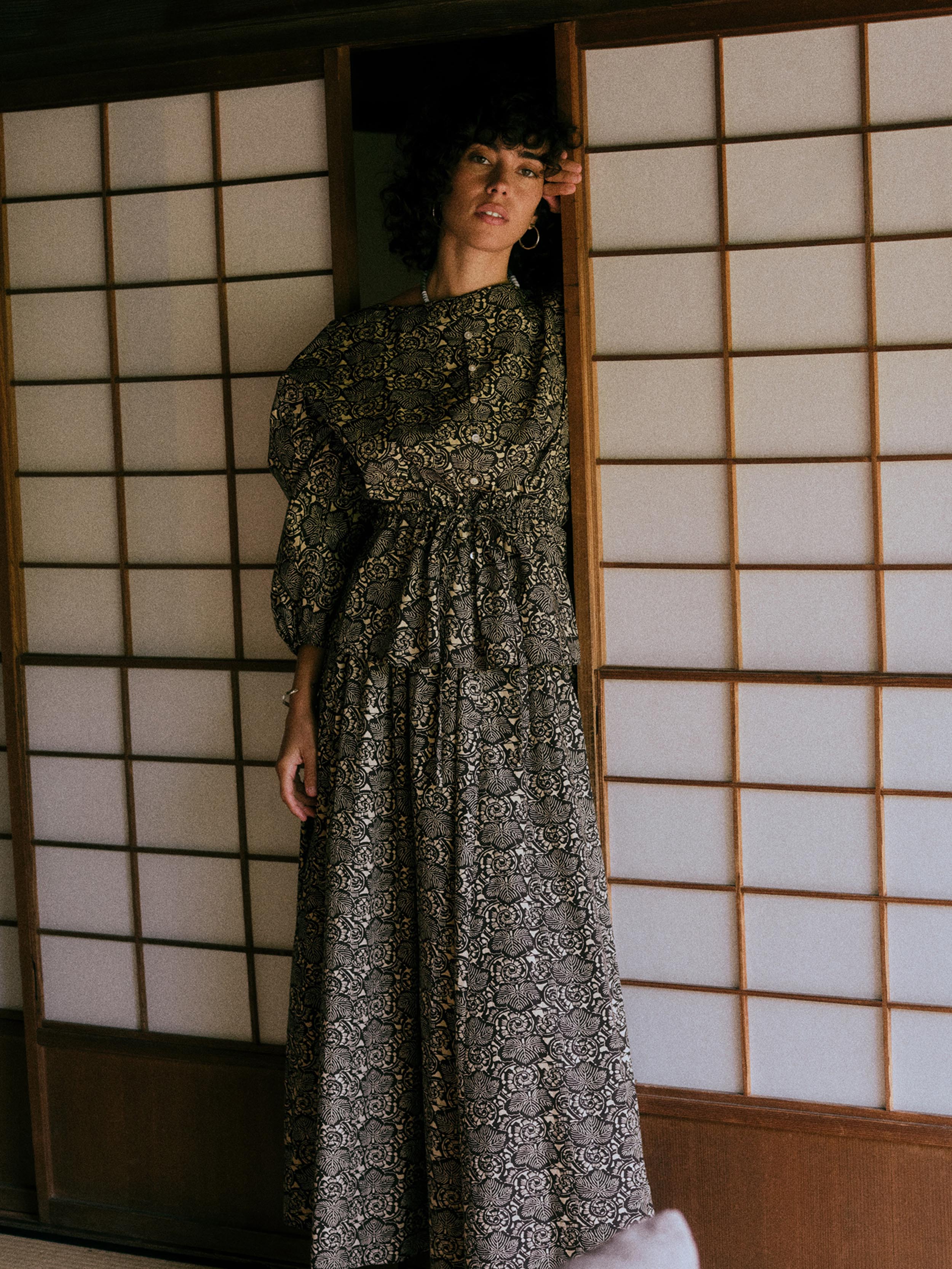 A woman wearing a long-sleeved, floor-length black and gray patterned dress stands in a doorway with traditional Japanese shoji screens behind her.