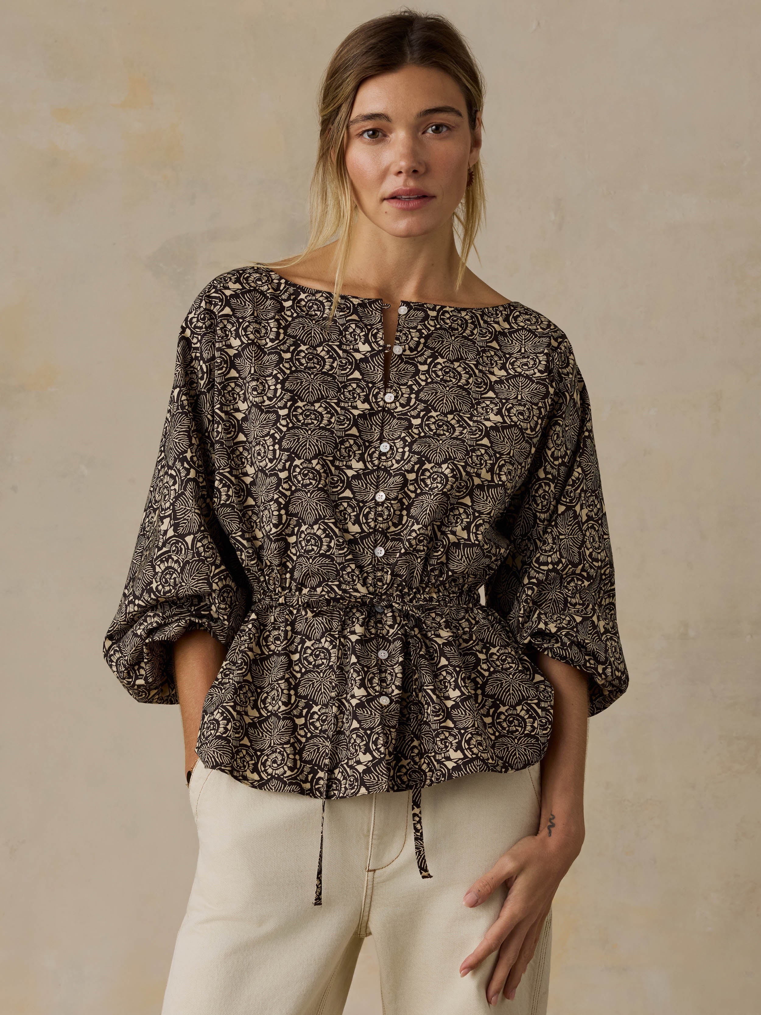 Woman wearing a black patterned blouse and white jeans against a beige background