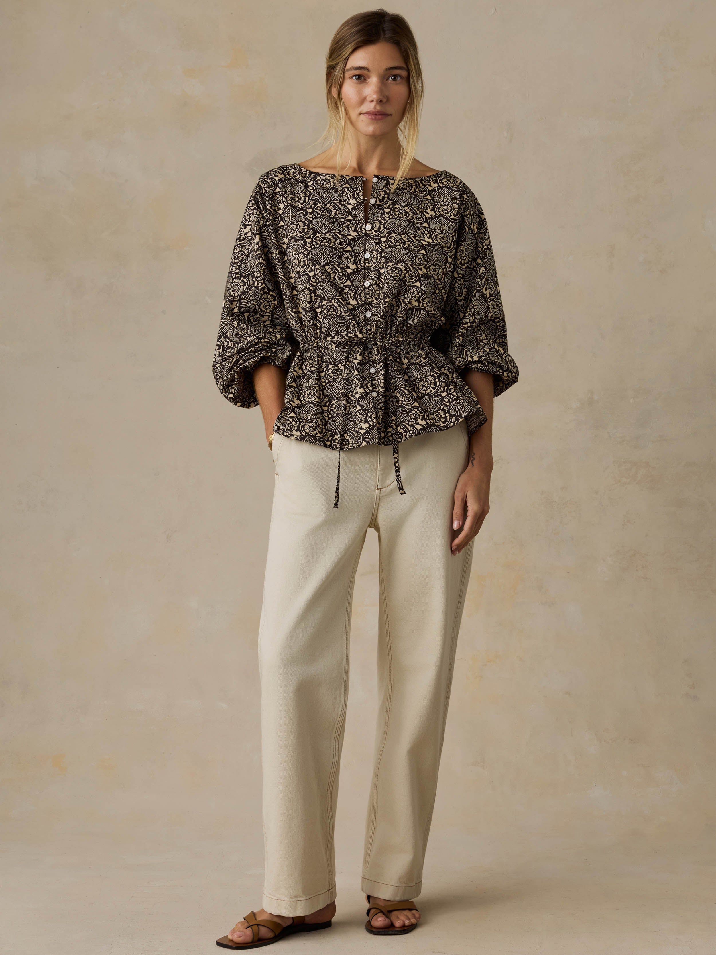Woman wearing a black patterned blouse and white jeans against a beige background