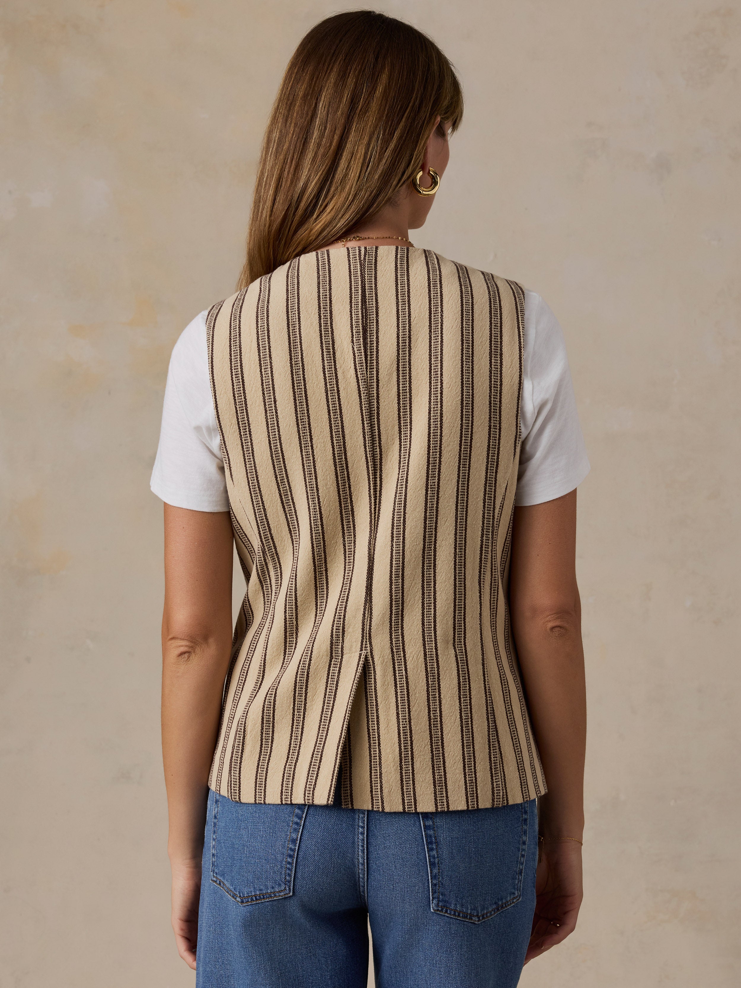 A woman viewed from the back wearing a sleeveless beige vest with vertical dark brown stripes over a white t-shirt and blue jeans, set against a textured beige background.