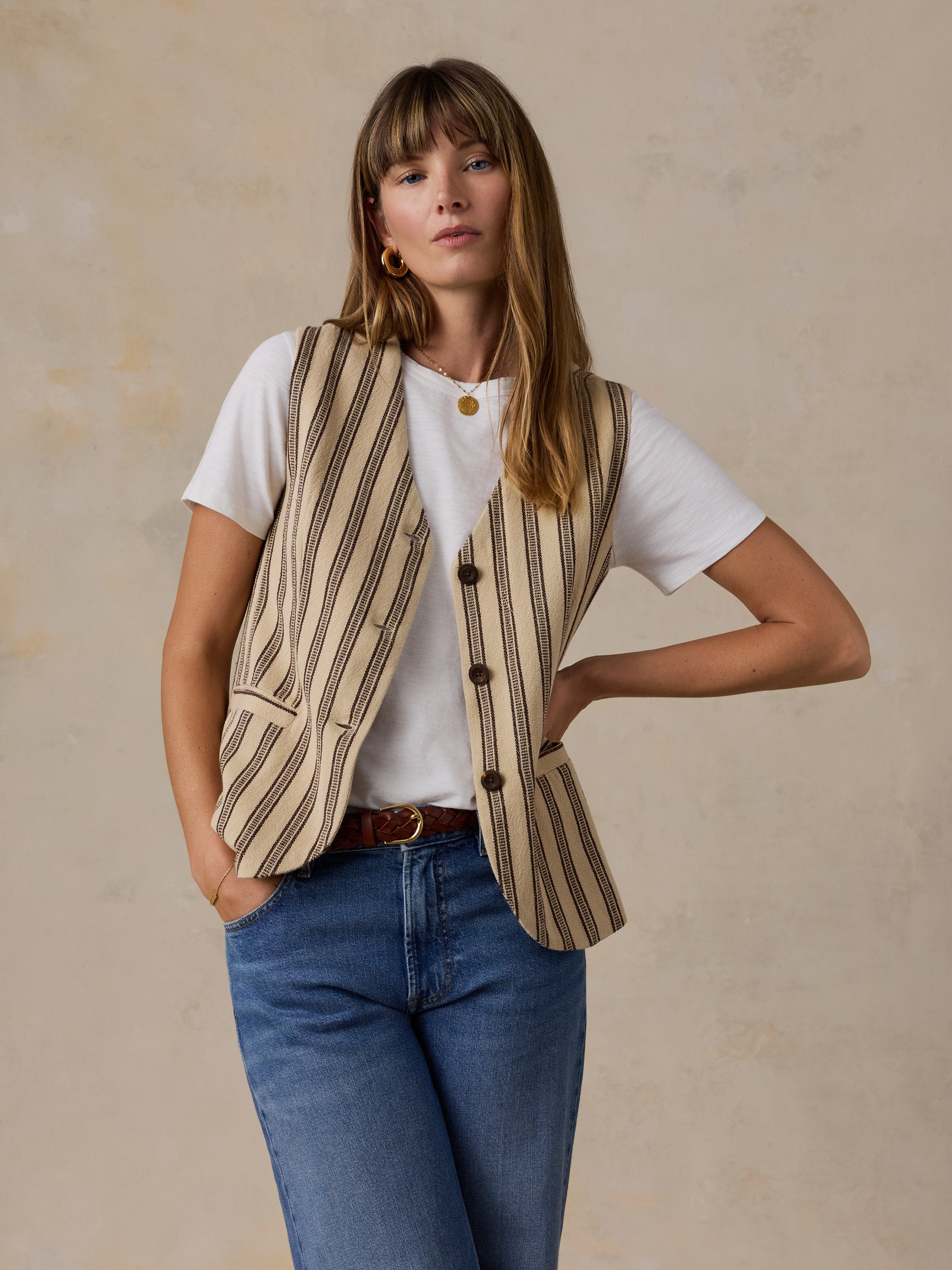 A woman modeling a beige and brown vertically striped button-up blazer vest over a white t-shirt and blue jeans, standing against a textured beige wall.