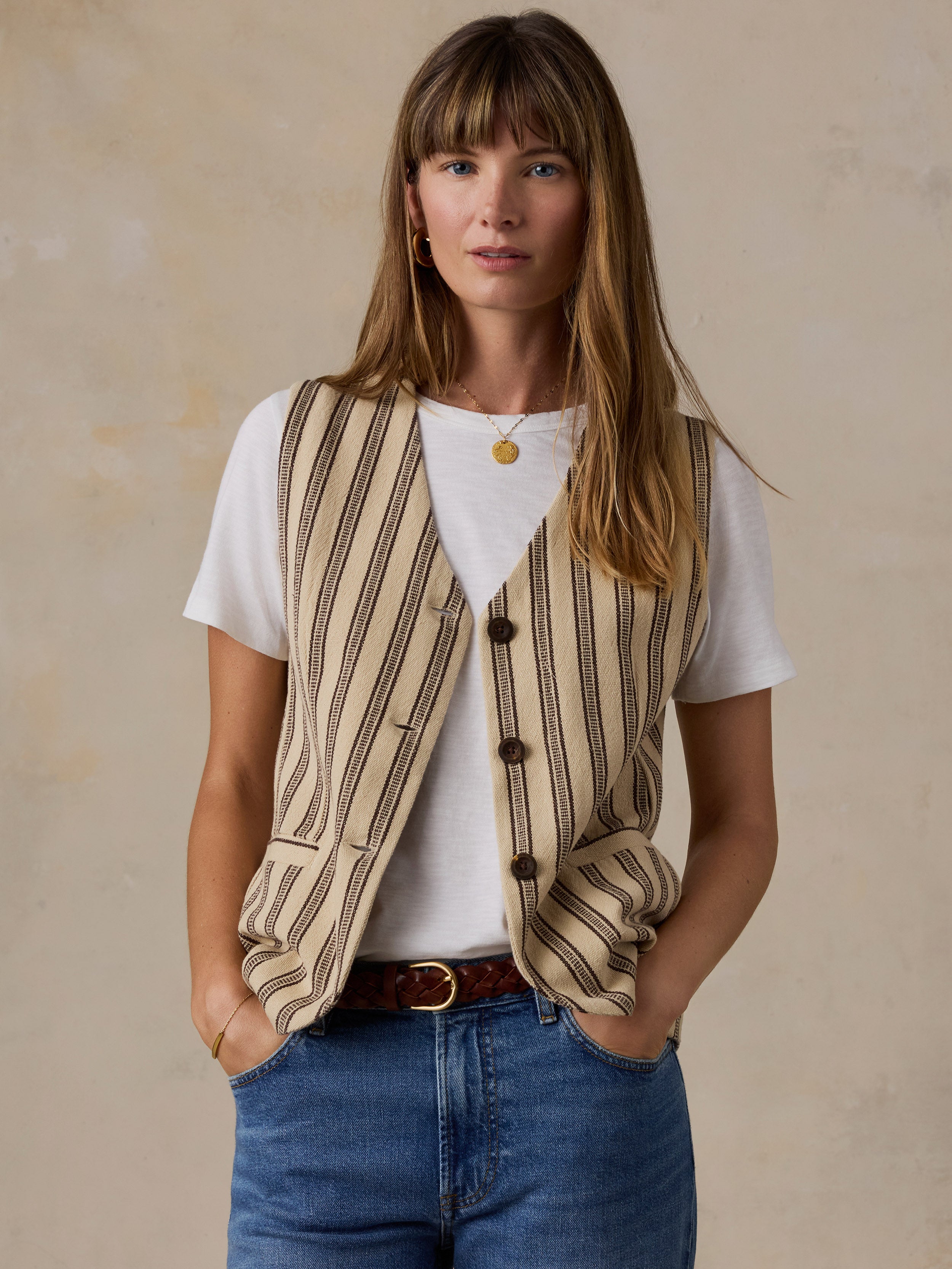 A woman wearing a beige and brown vertically striped knit vest over a white t-shirt, paired with blue jeans and a brown belt, standing against a textured beige wall.