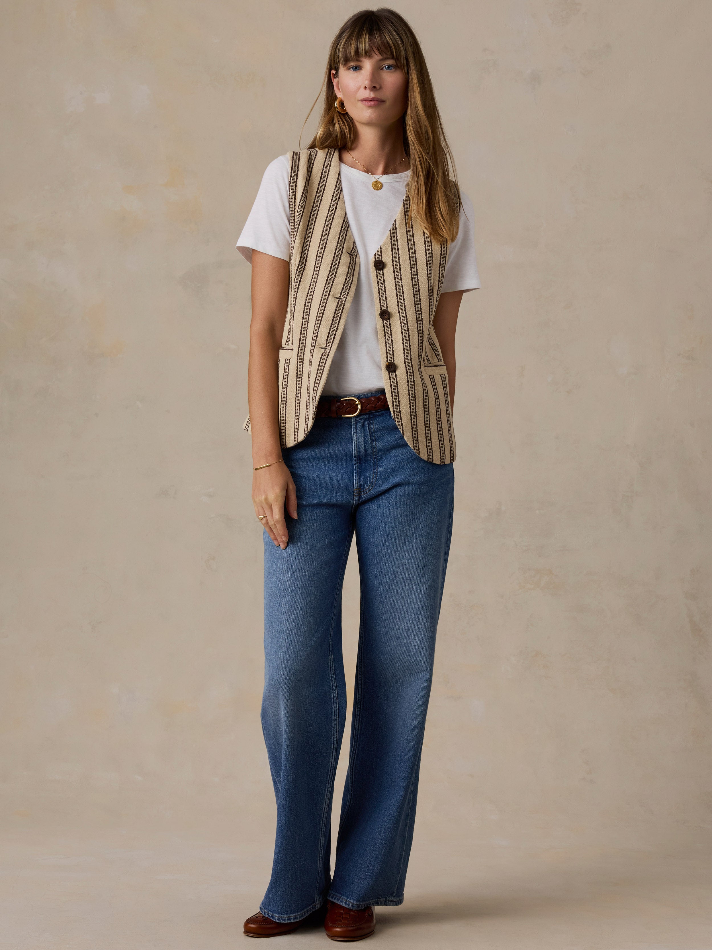 A woman stands against a textured beige studio backdrop wearing a cream and tan vertically striped vest over a white T-shirt, blue jeans, and brown shoes.