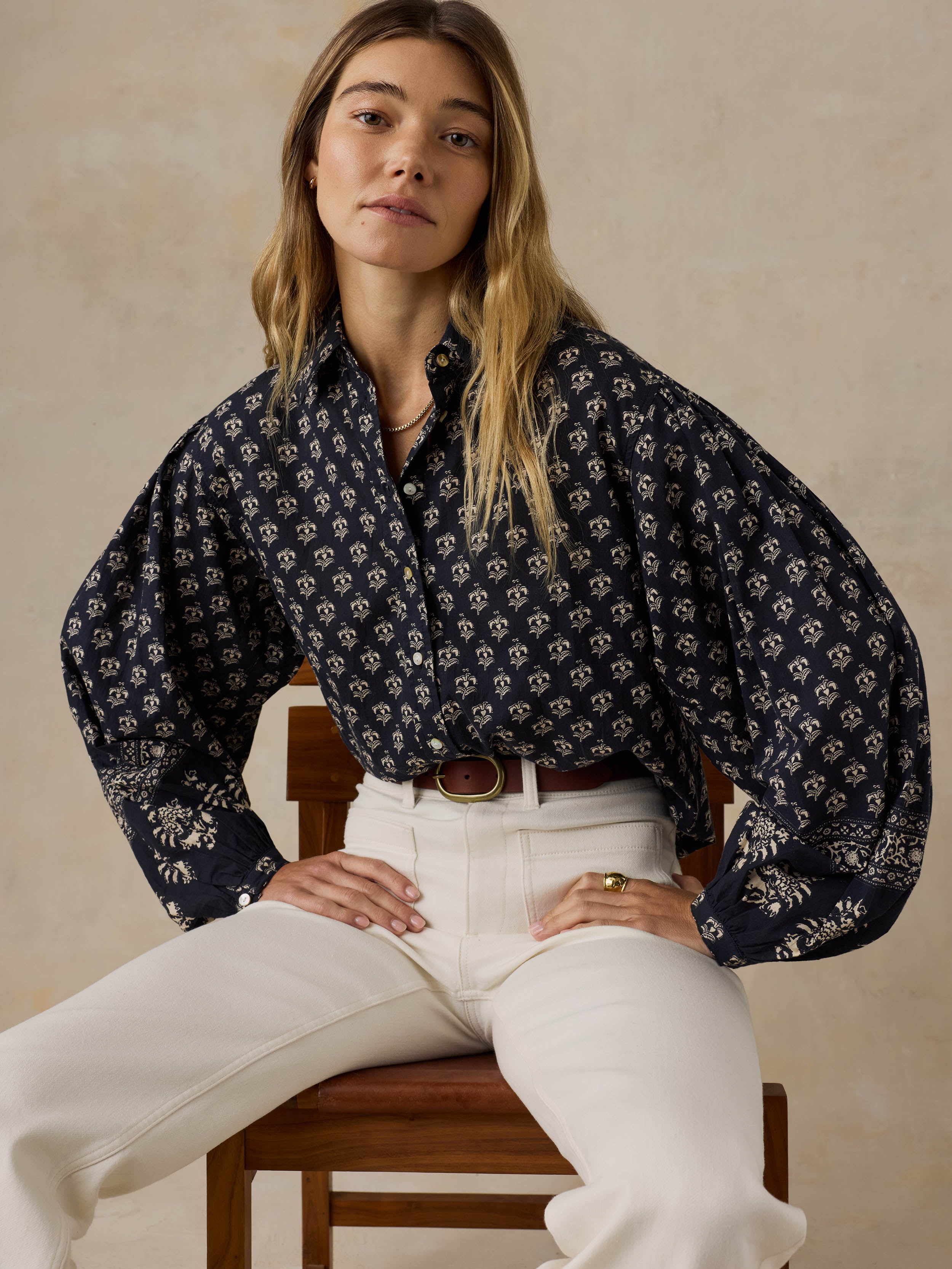 Woman sitting on a wooden chair wearing a dark patterned blouse and white pants against a beige background