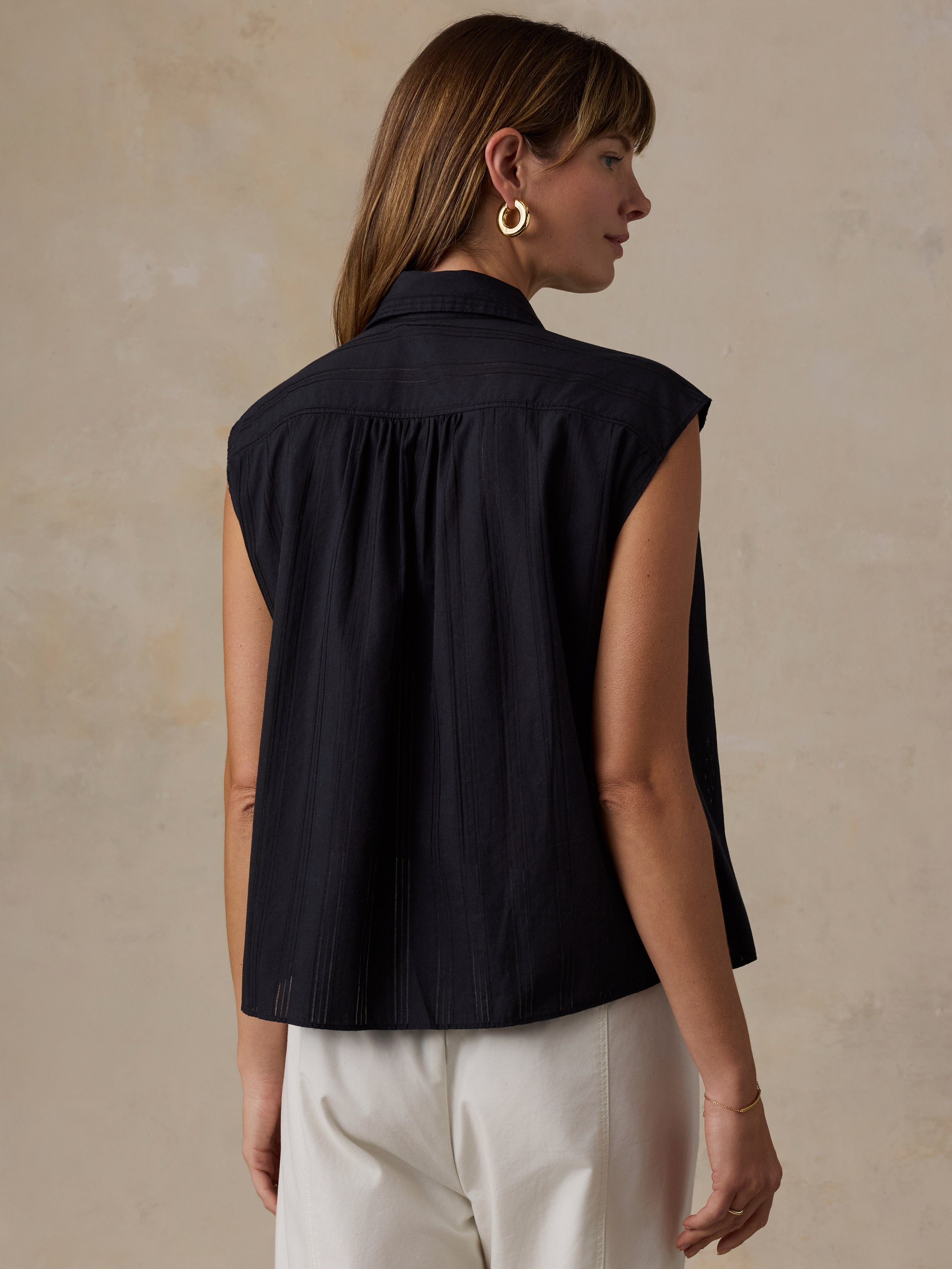 A woman shown from the back wearing a sleeveless black top with a gathered neckline and white pants, standing against a textured beige wall.