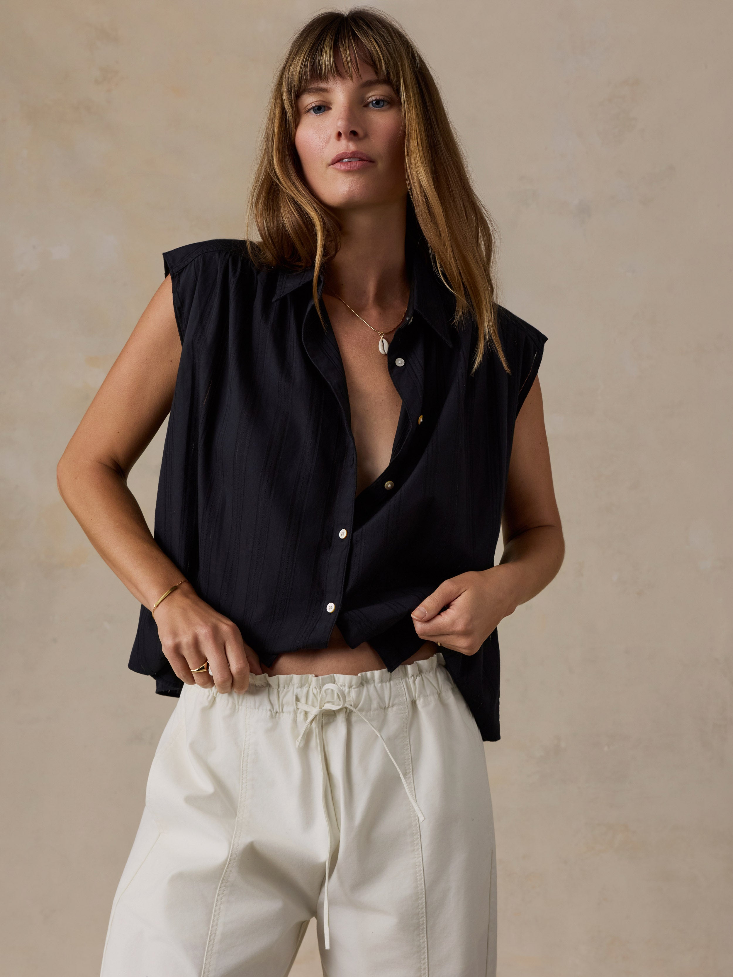 A woman wearing a sleeveless black button-up shirt and cream drawstring trousers, standing against a textured beige wall.