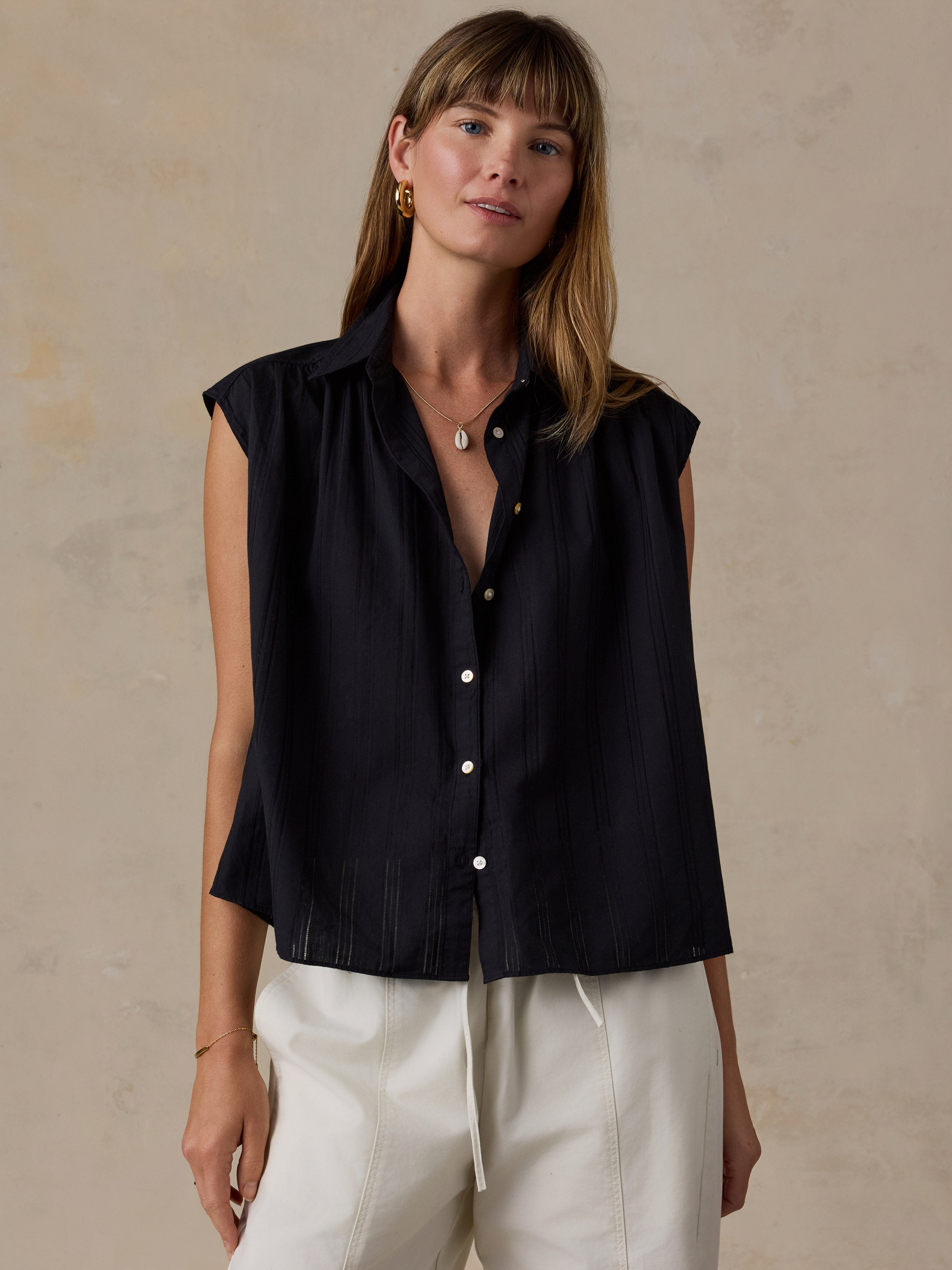 A woman wearing a sleeveless black button-front shirt with white drawstring pants, standing against a textured beige wall.