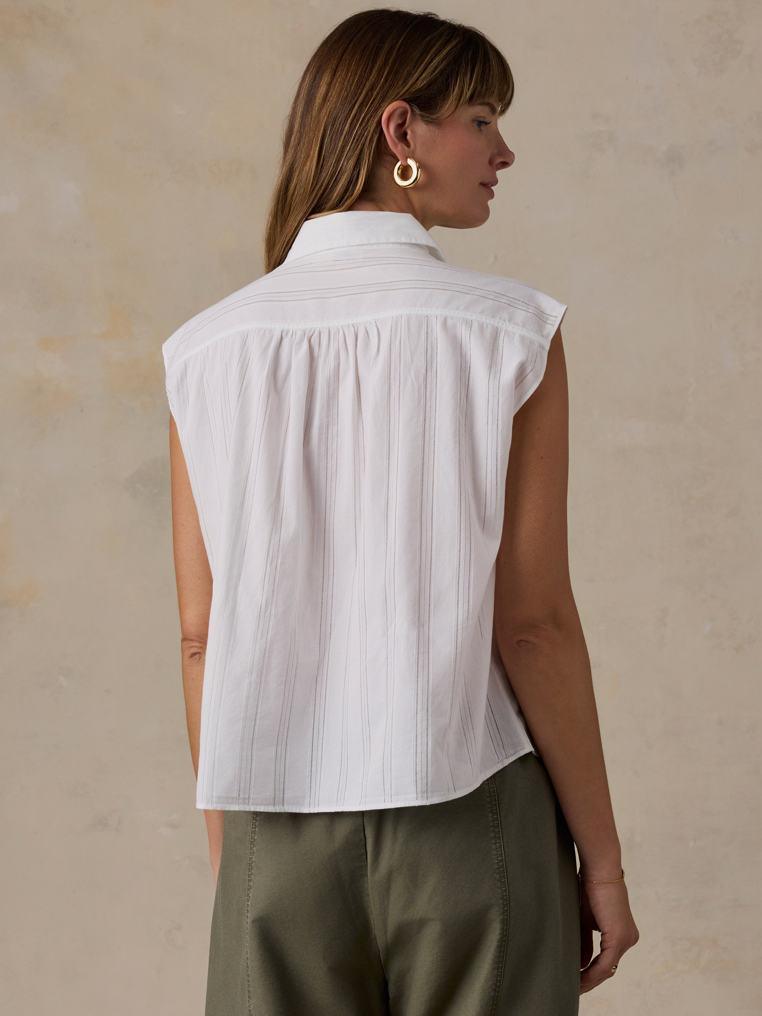 A woman shown from the back wearing a white sleeveless collared blouse with subtle vertical texture and pleated detailing, paired with olive green pants, against a textured beige wall.
