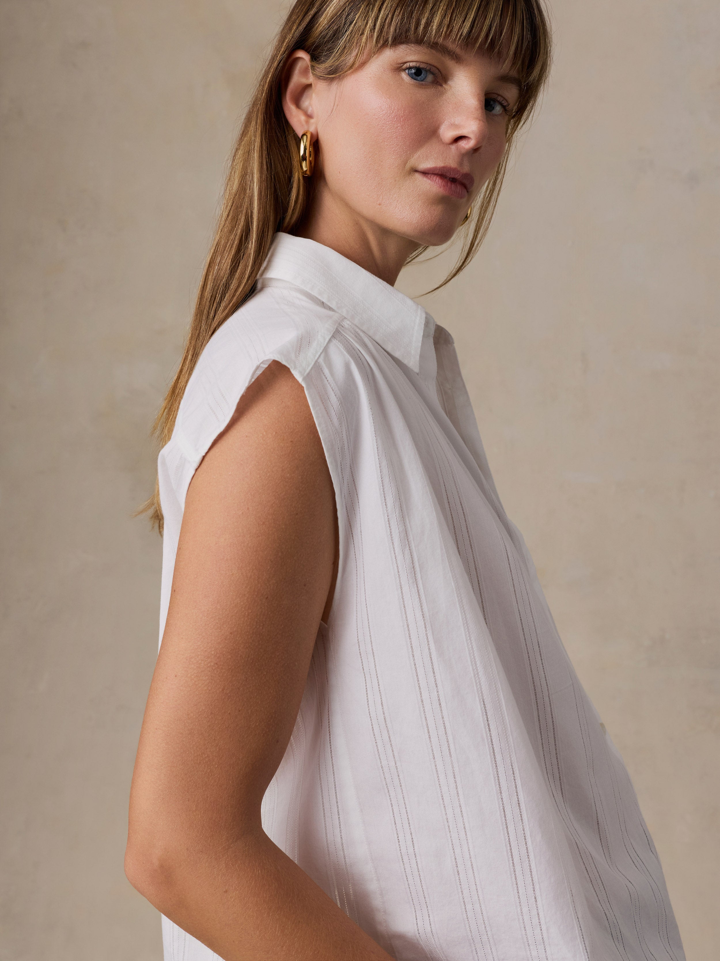 A woman wearing a sleeveless white collared blouse with a subtle vertical pleated texture, photographed against a warm beige textured background.