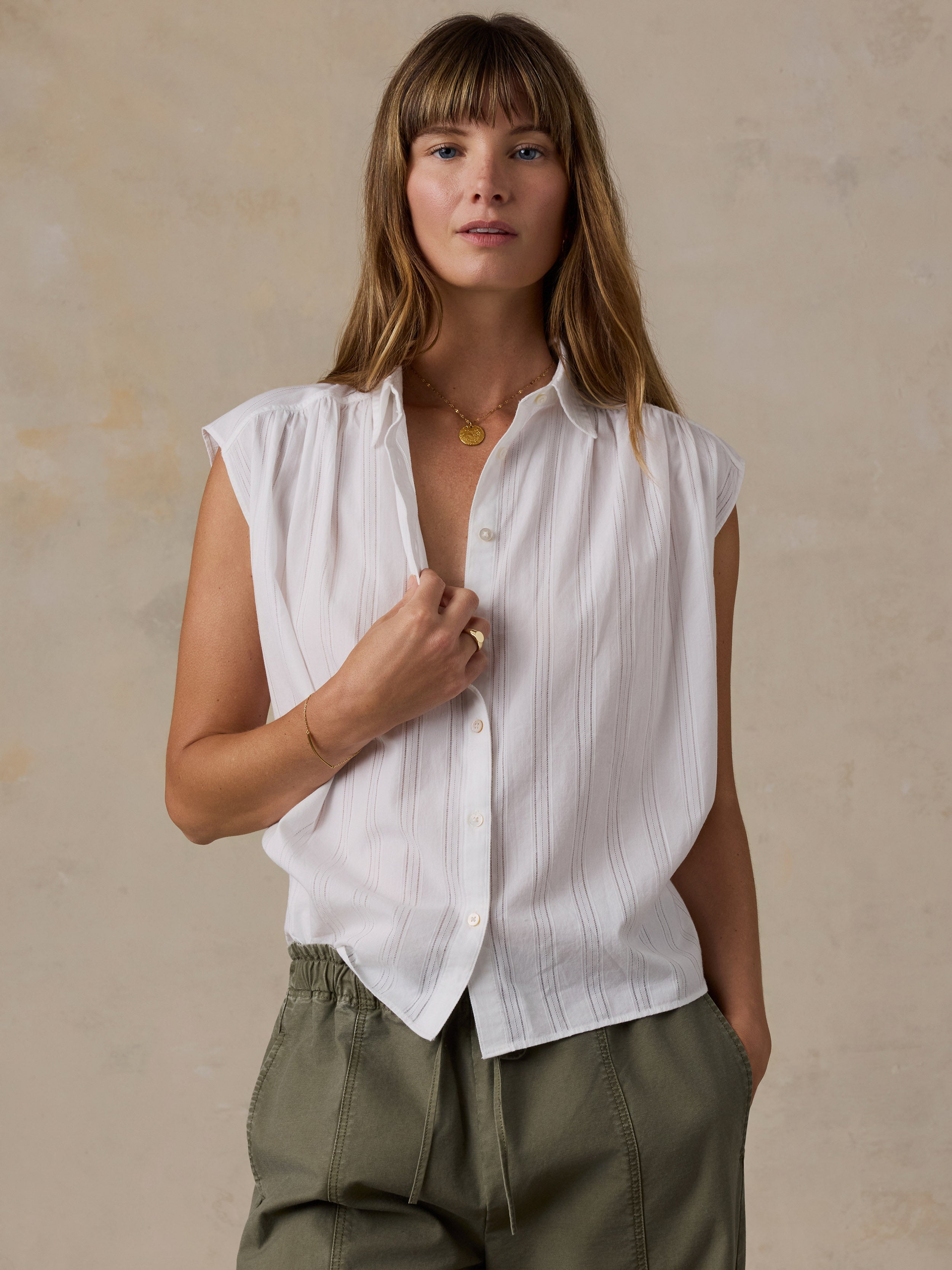A woman wearing a white sleeveless button-up shirt with subtle vertical pinstripes and olive green pants, standing against a textured beige studio background.