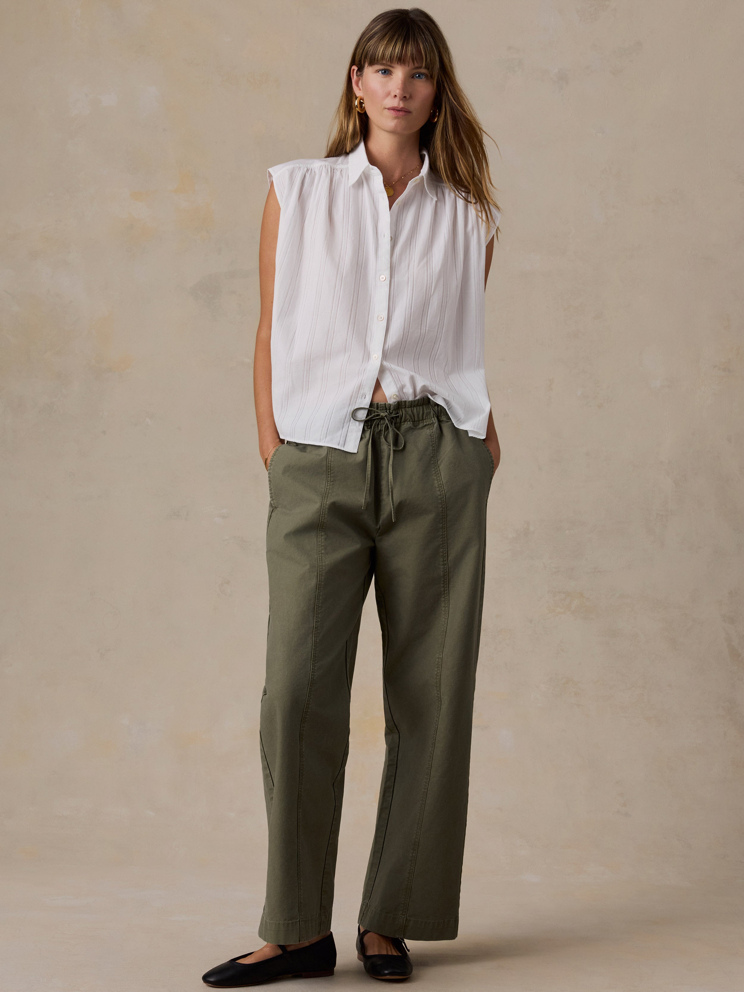 A woman stands against a textured beige studio backdrop wearing a sleeveless white button-up shirt and olive green drawstring pants with black flats.