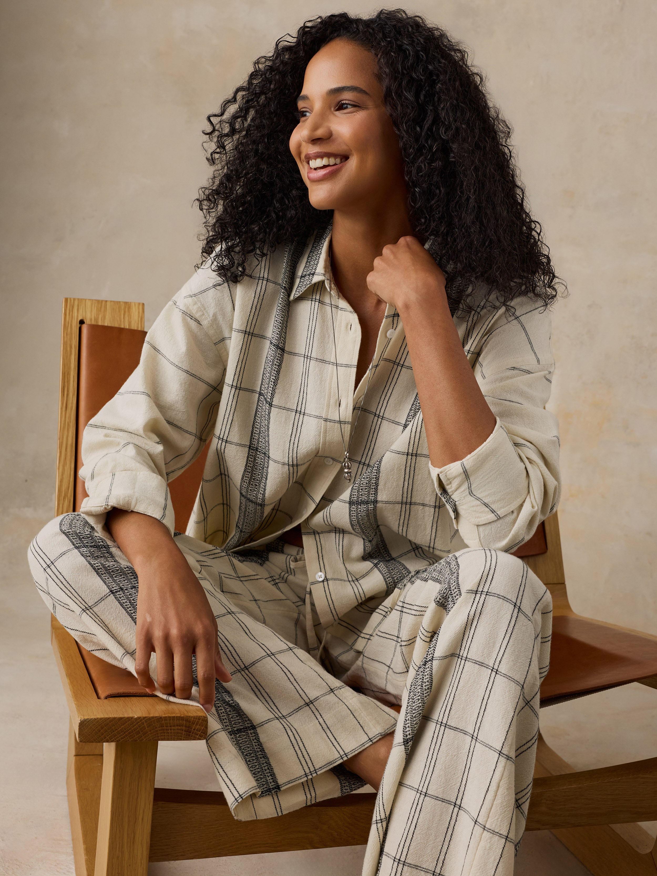 Woman sitting on a wooden chair wearing a white and black plaid shirt and matching pants against a beige background