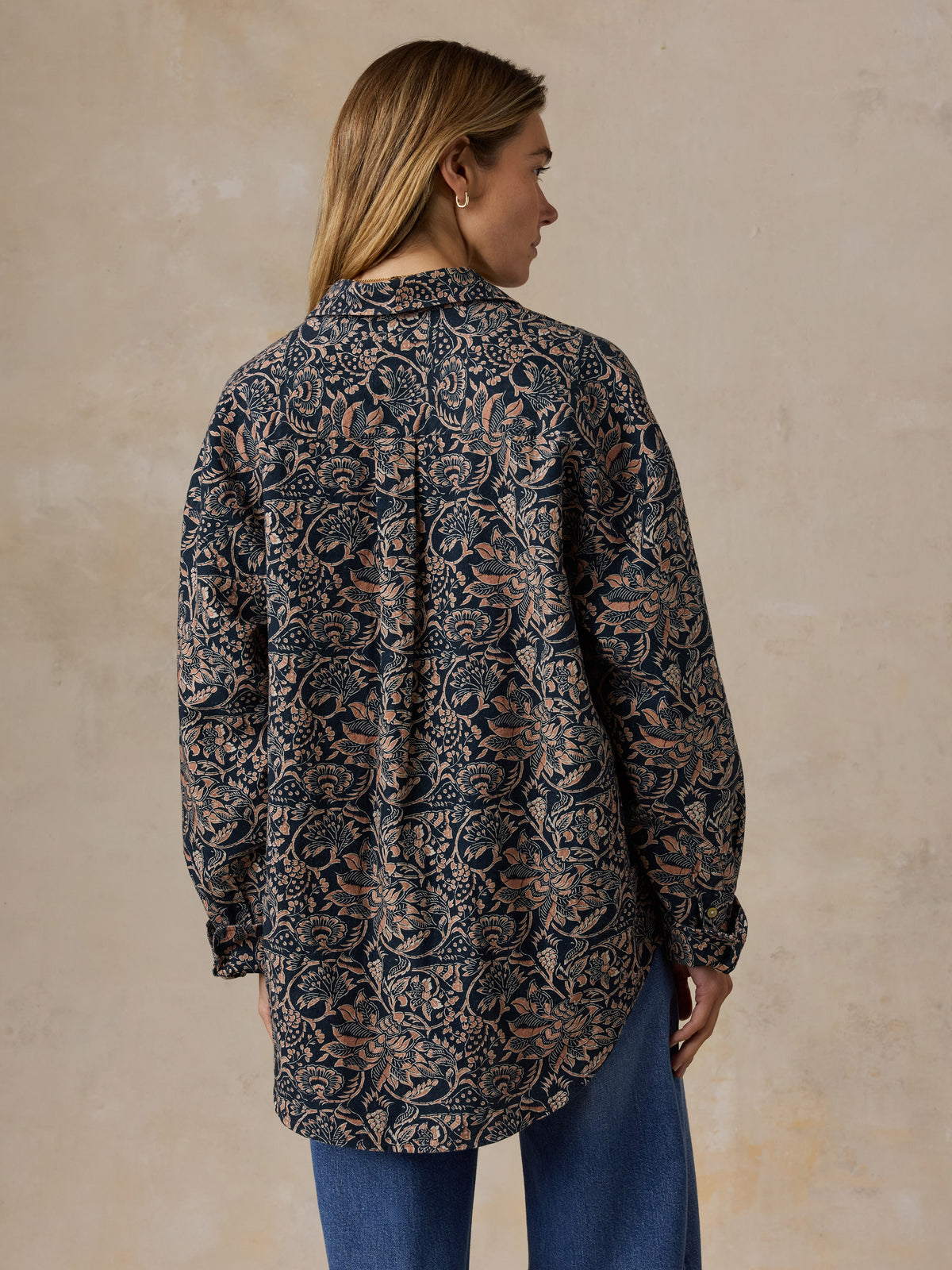 A woman shown from the back wearing a long-sleeve button-up shirt with an intricate dark navy and brown floral damask pattern, paired with blue jeans, set against a textured beige wall., image 4 of 7
