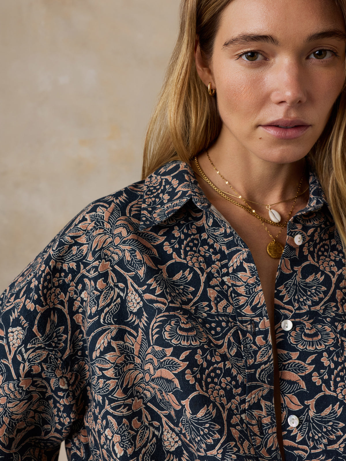 A close-up of a woman wearing a dark navy button-up shirt with a detailed beige and brown floral print, styled with layered gold necklaces against a neutral textured beige background., image 5 of 7
