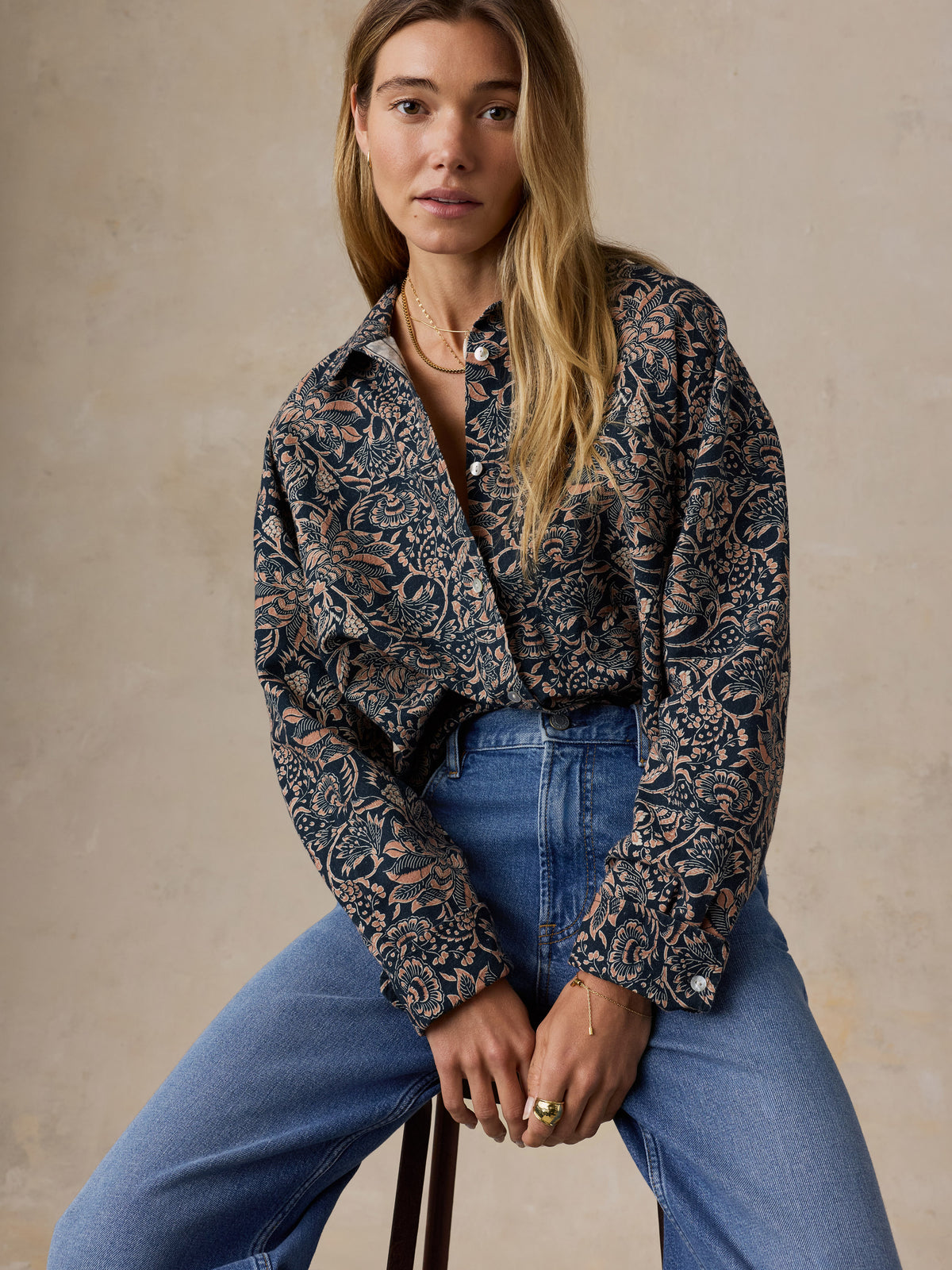 A woman wearing a long-sleeve button-up shirt with an intricate dark blue and beige floral pattern, tucked into blue jeans, sitting against a textured beige wall., image 6 of 7
