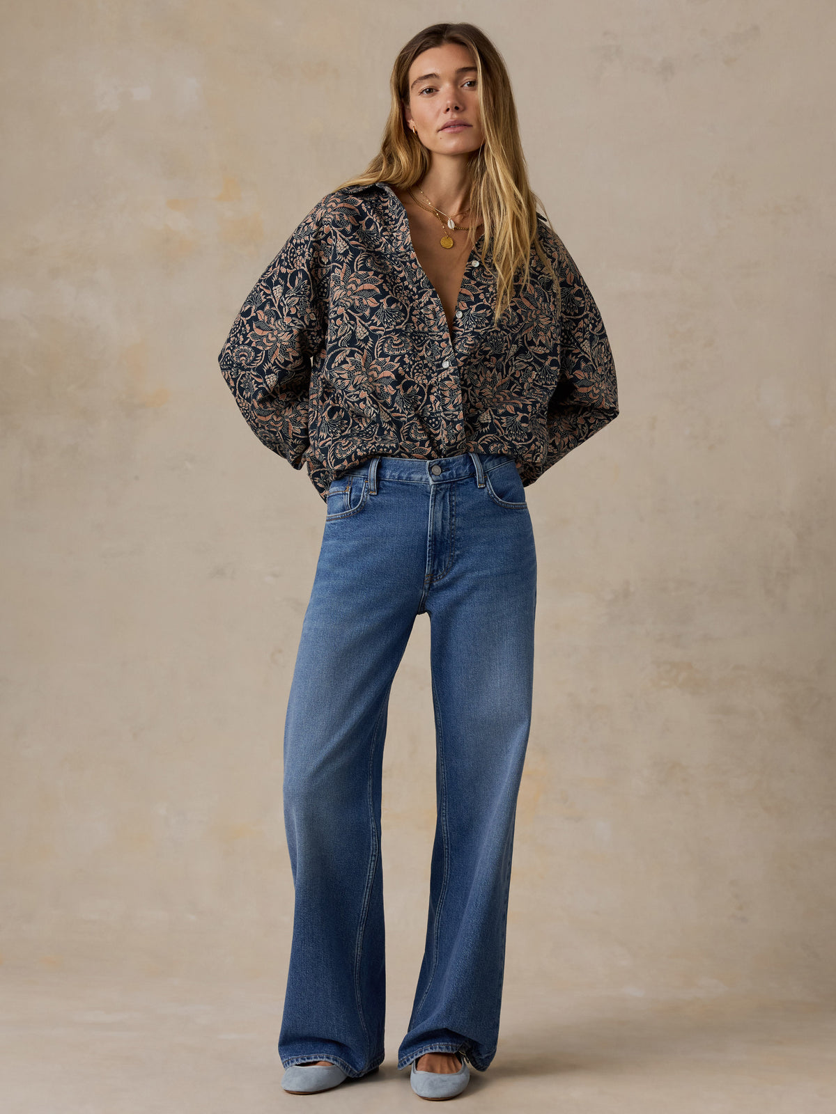 A woman stands against a textured beige studio backdrop wearing a dark patterned button-up blouse with wide sleeves and blue wide-leg jeans., image 7 of 7