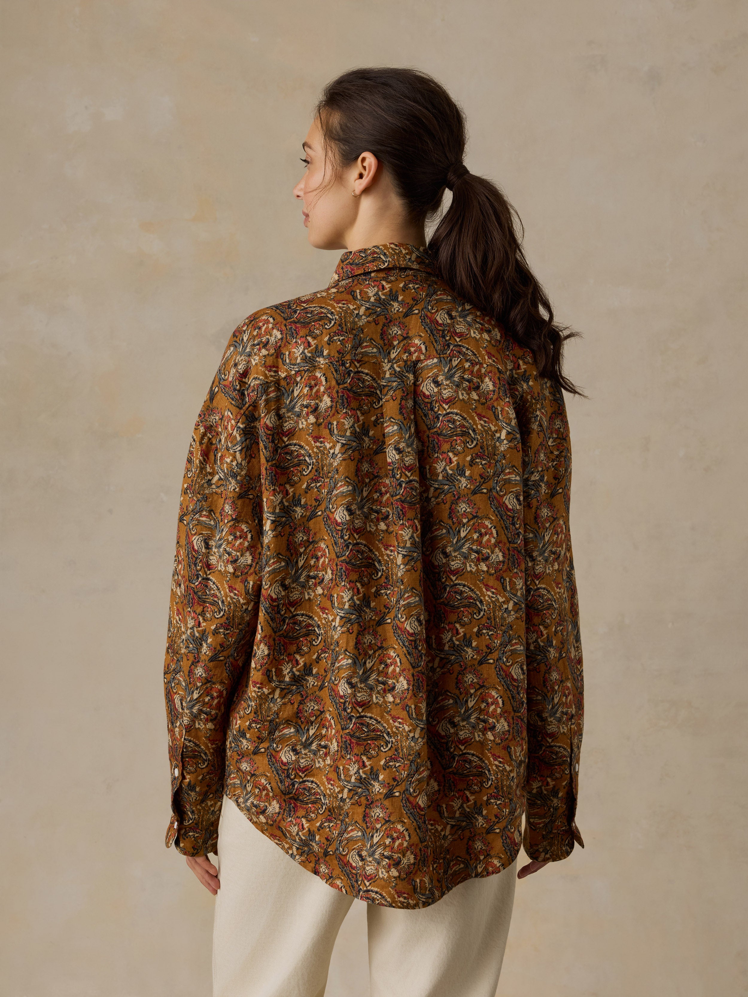 Woman wearing an orange floral printed button down shirt with white pants against a beige background