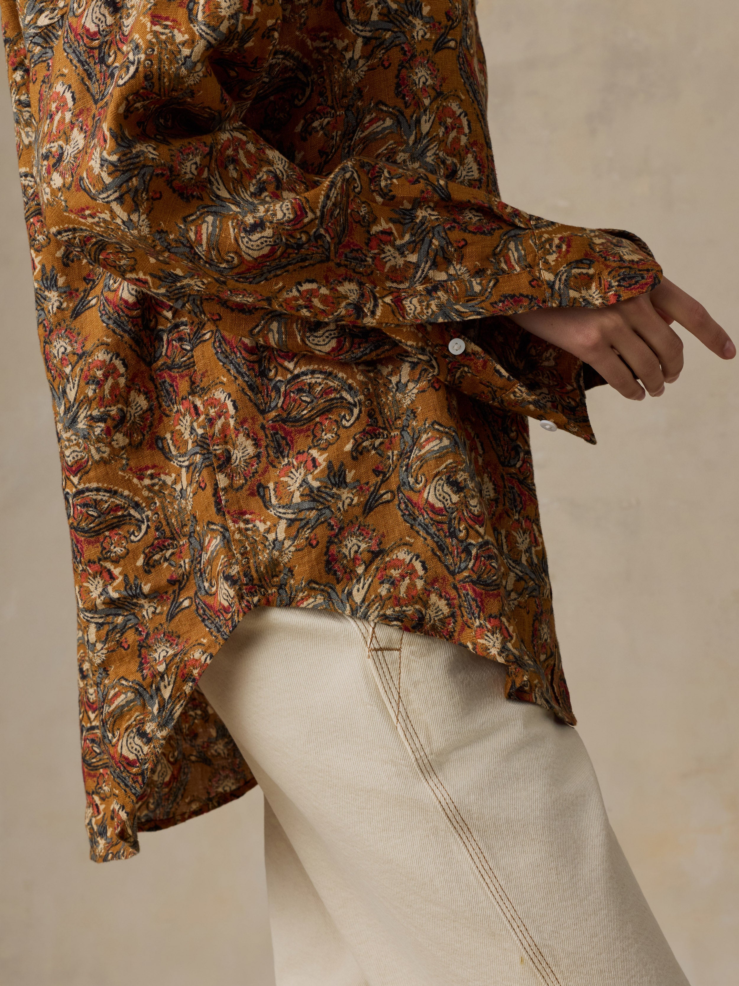 Woman wearing an orange floral printed button down shirt with white pants against a beige background