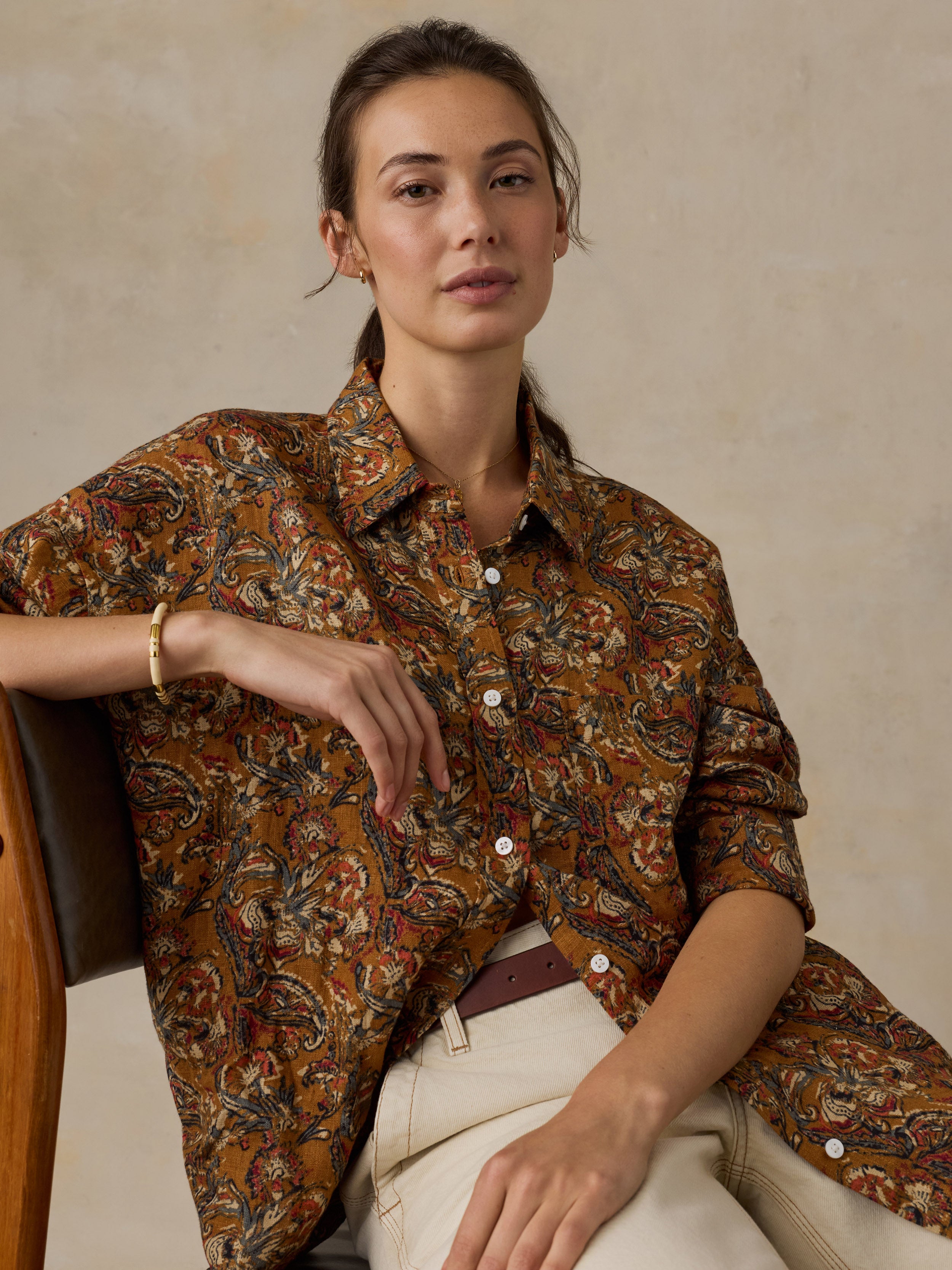 Woman sitting on a wooden chair wearing an orange floral printed button down shirt with white pants against a beige background
