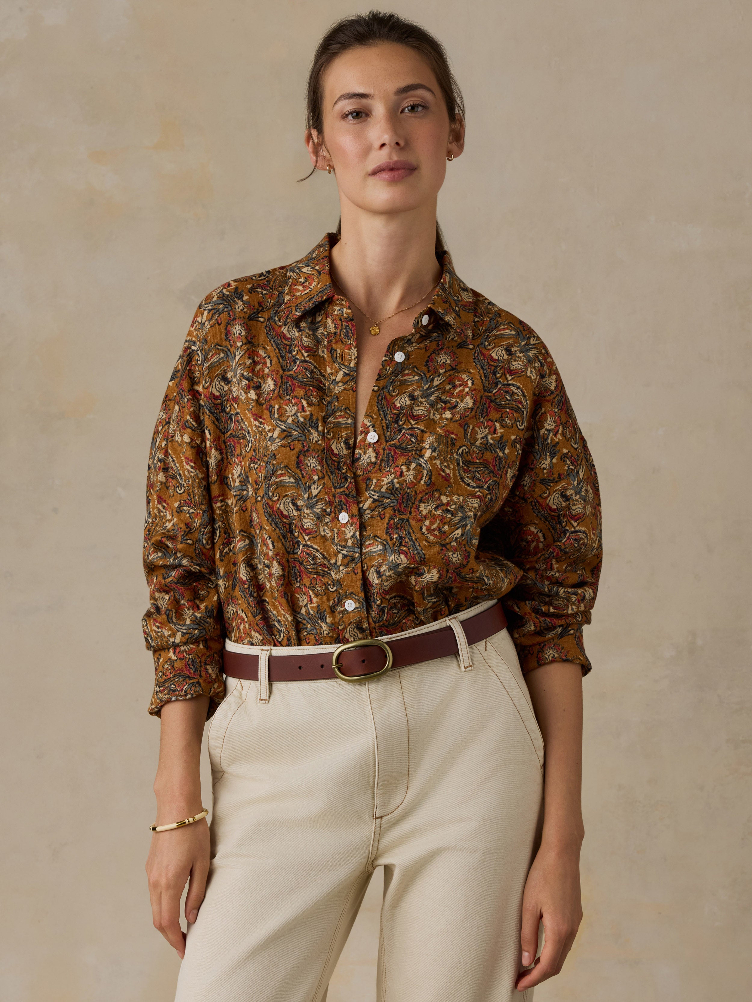 Woman wearing an orange floral printed button down shirt with white pants against a beige background