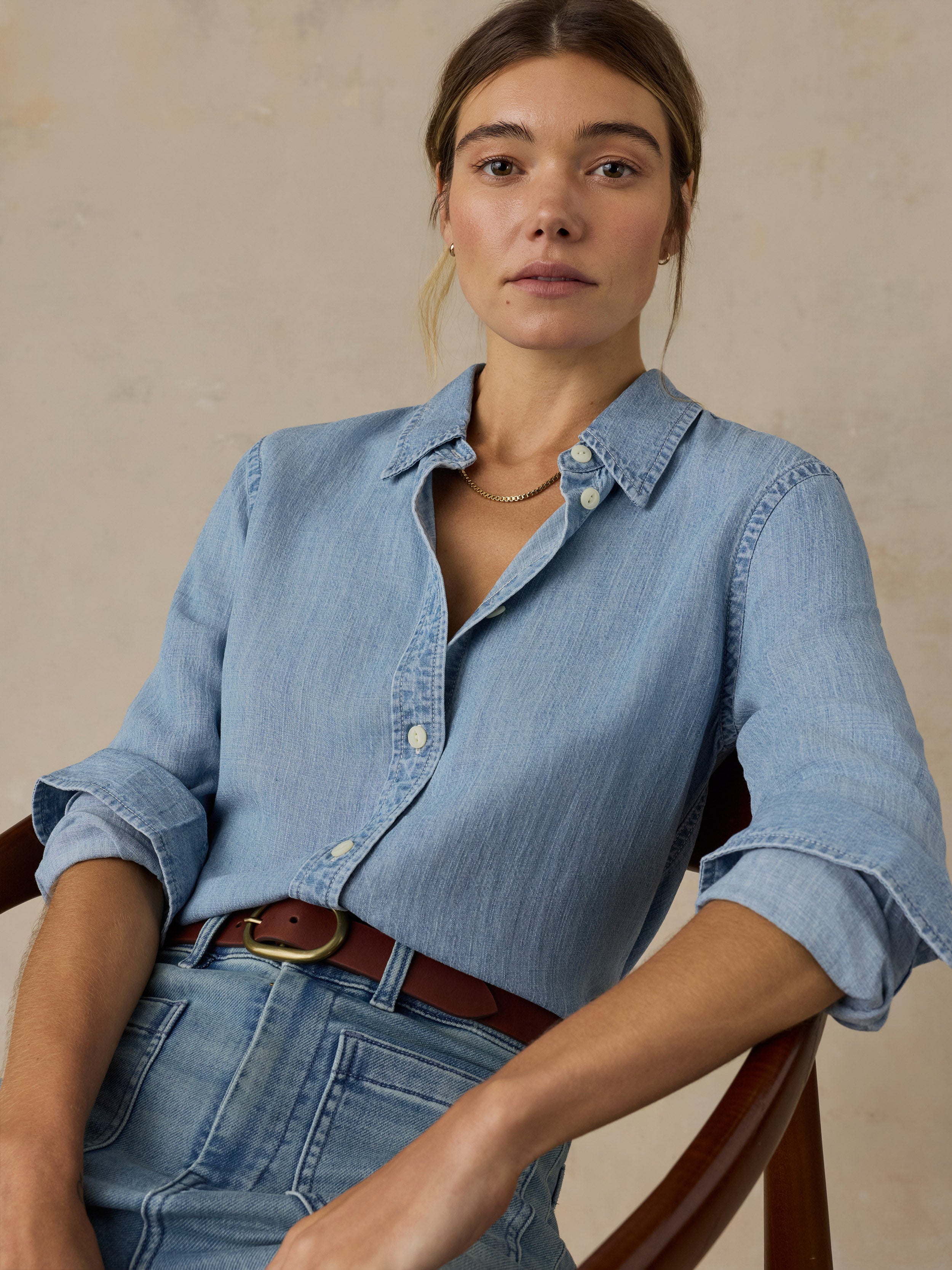 Woman sitting on a wooden chair wearing a chambray button down shirt with blue jeans against a beige background
