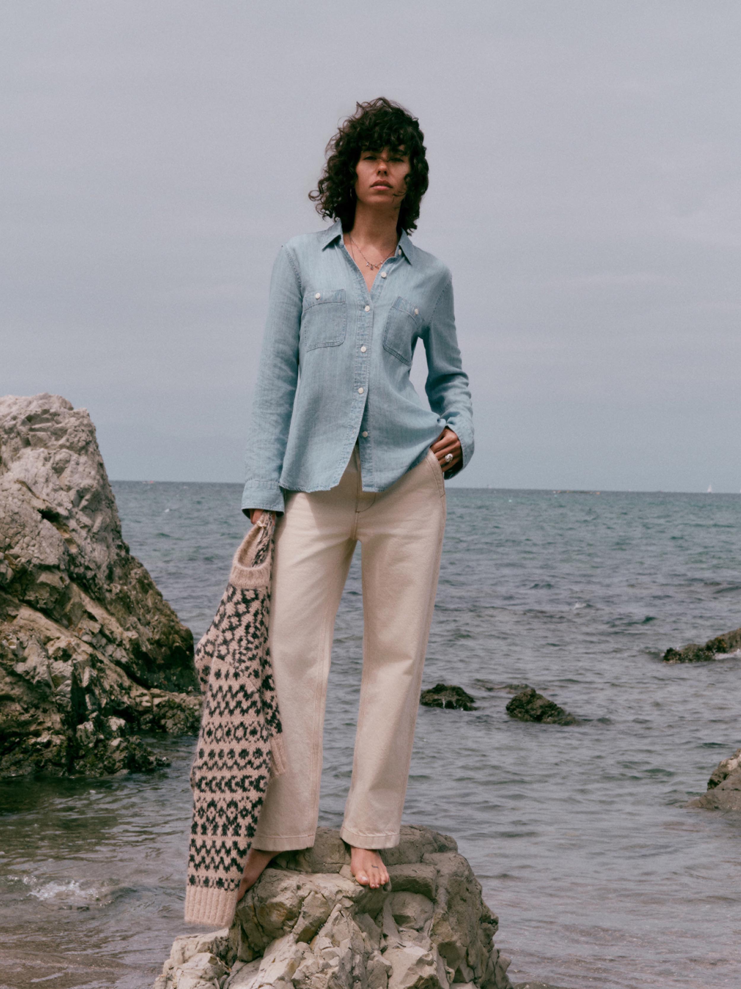 A woman stands barefoot on a rocky shoreline wearing a light blue button-up shirt, cream wide-leg pants, and holding a patterned knit cardigan against an overcast sea background.