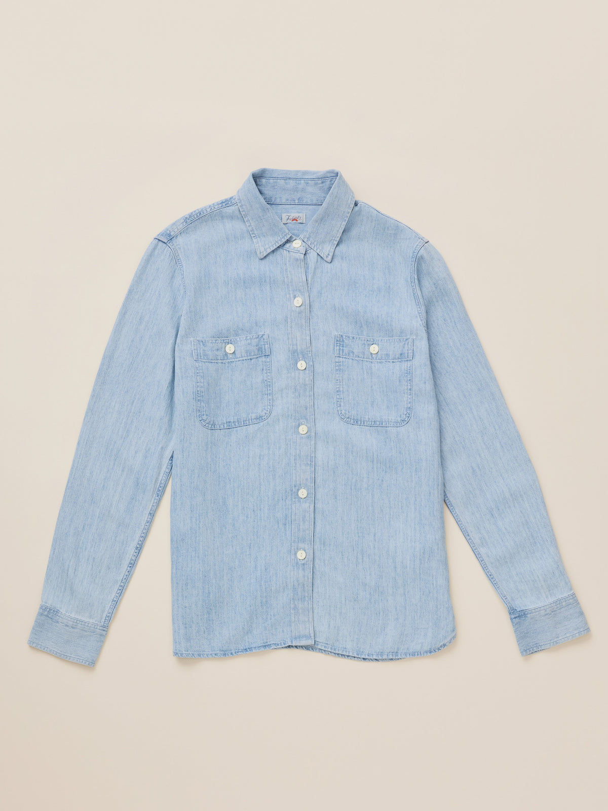 A light blue denim long-sleeve button-up shirt with two chest pockets, laid flat on a warm beige background.