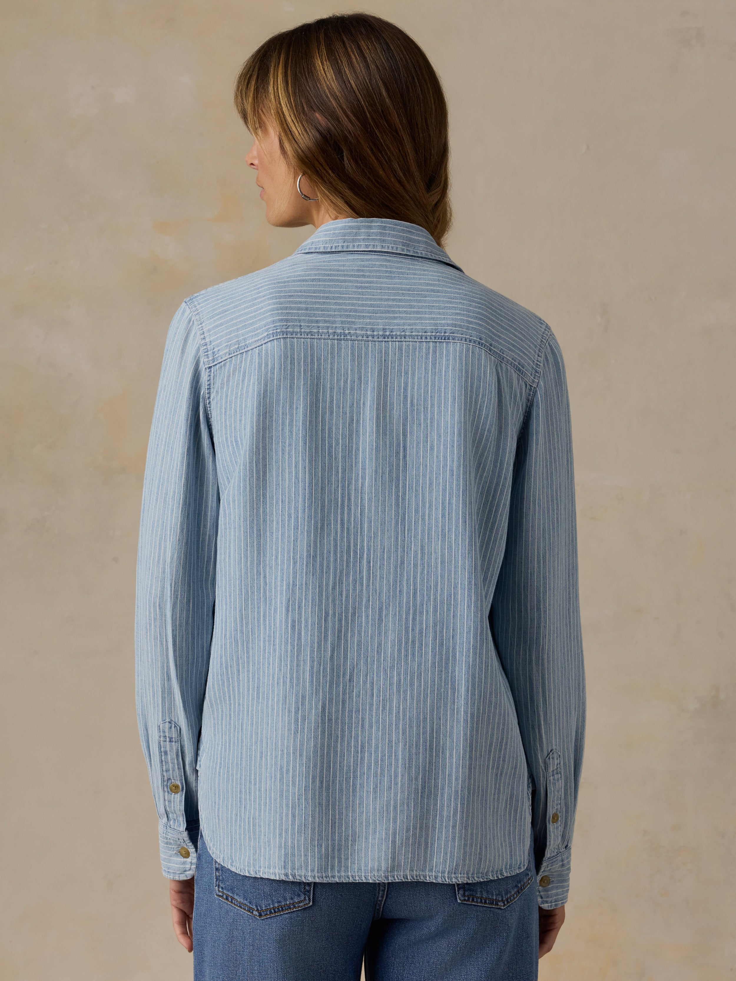 Woman wearing a blue and white striped chambray button down shirt and blue jeans against a beige background