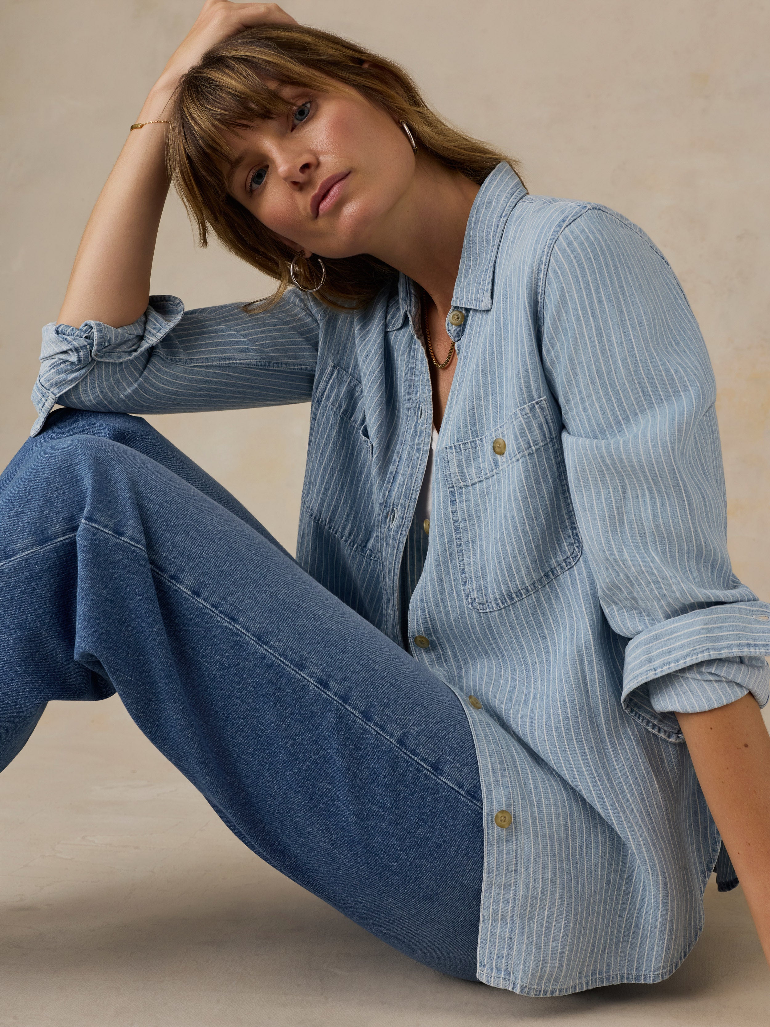 Woman wearing a blue and white striped chambray button down shirt over a white tank top and blue jeans against a beige background