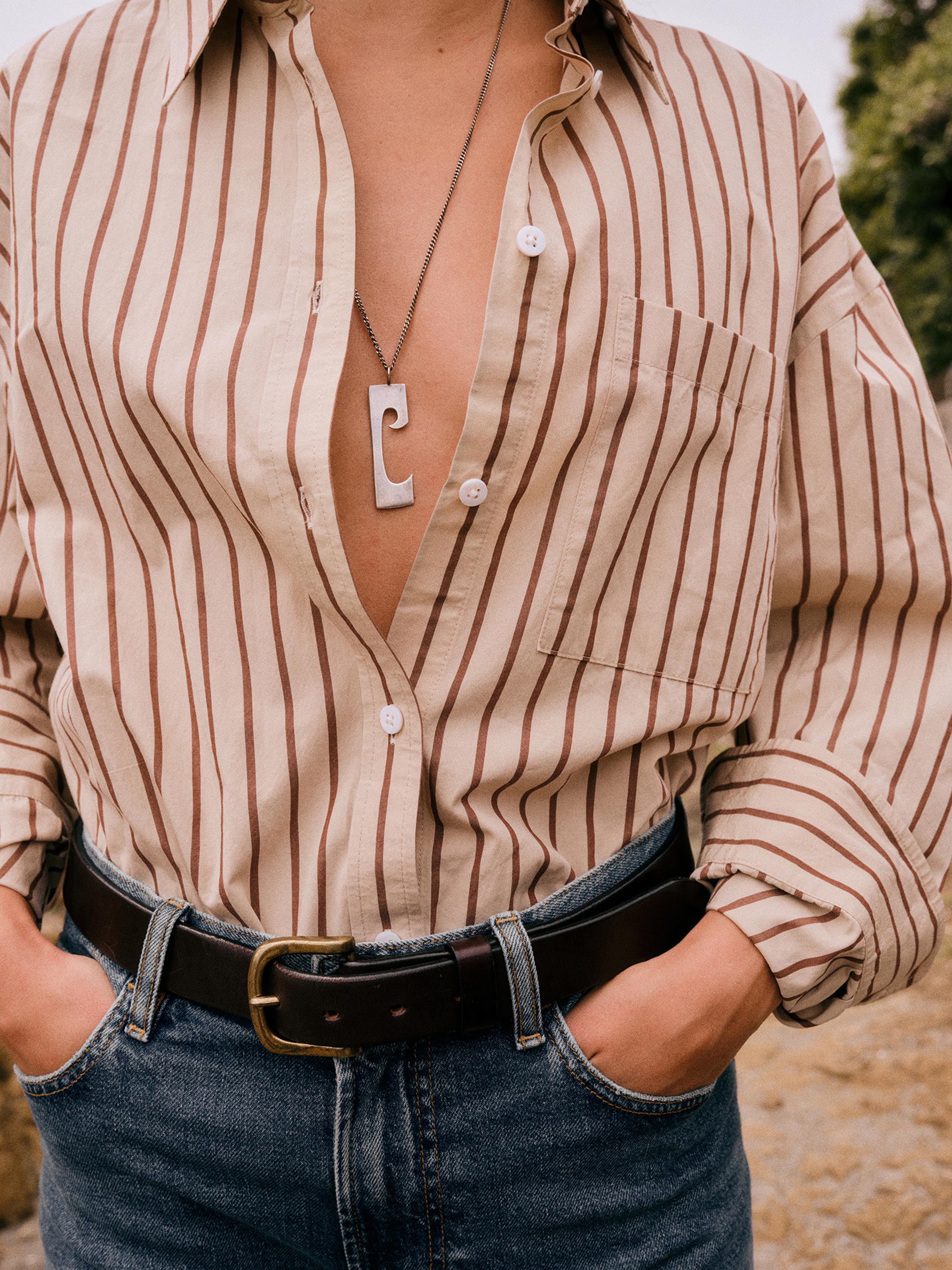 A woman wearing a beige button-up shirt with thin brown vertical stripes, tucked into blue jeans with a dark brown belt. The shirt is unbuttoned at the chest, showing a silver pendant necklace, and the background is a soft-focus outdoor setting.