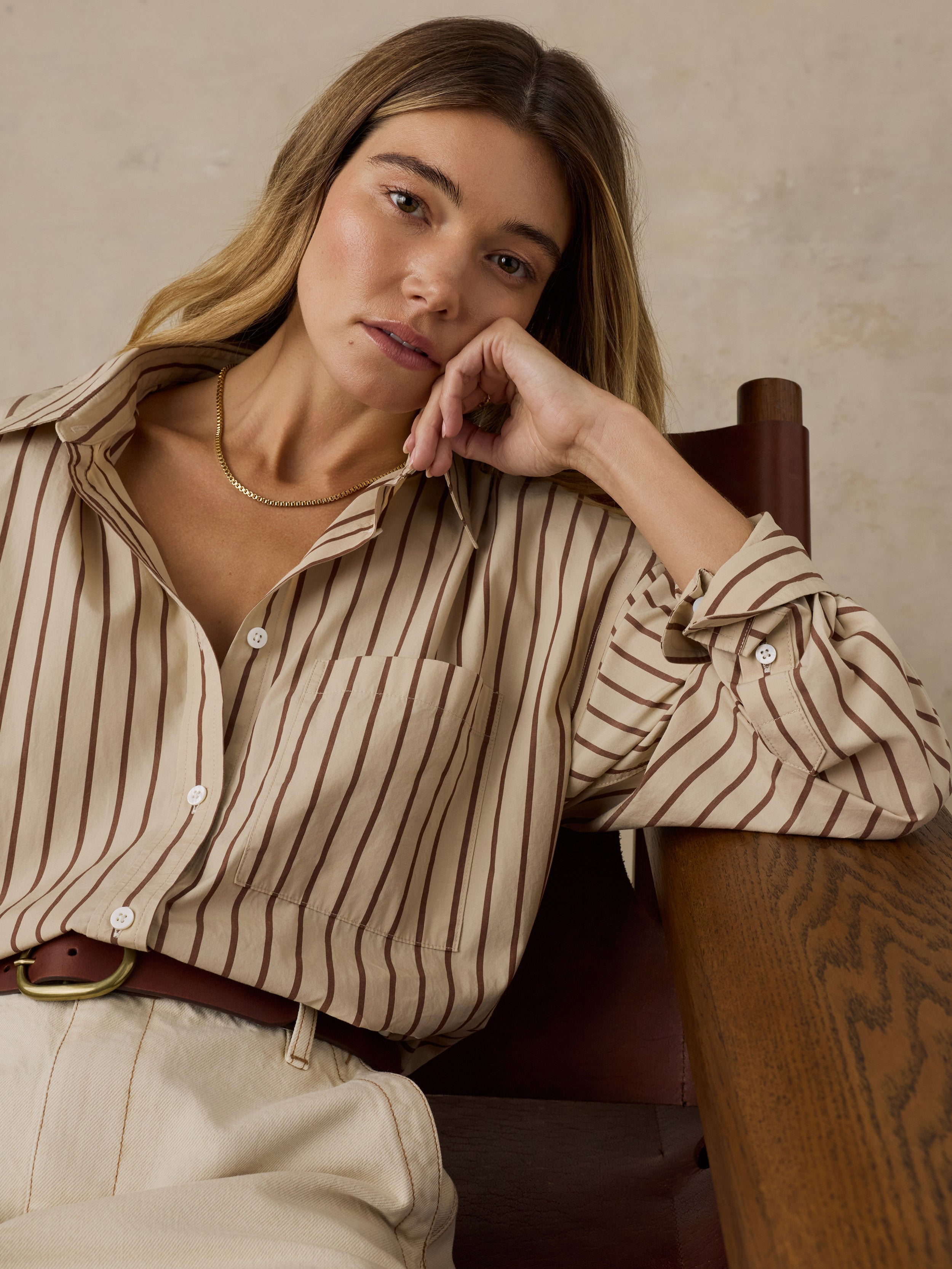 Woman sitting on a wooden chair wearing a beige and brown striped button down shirt against a beige background