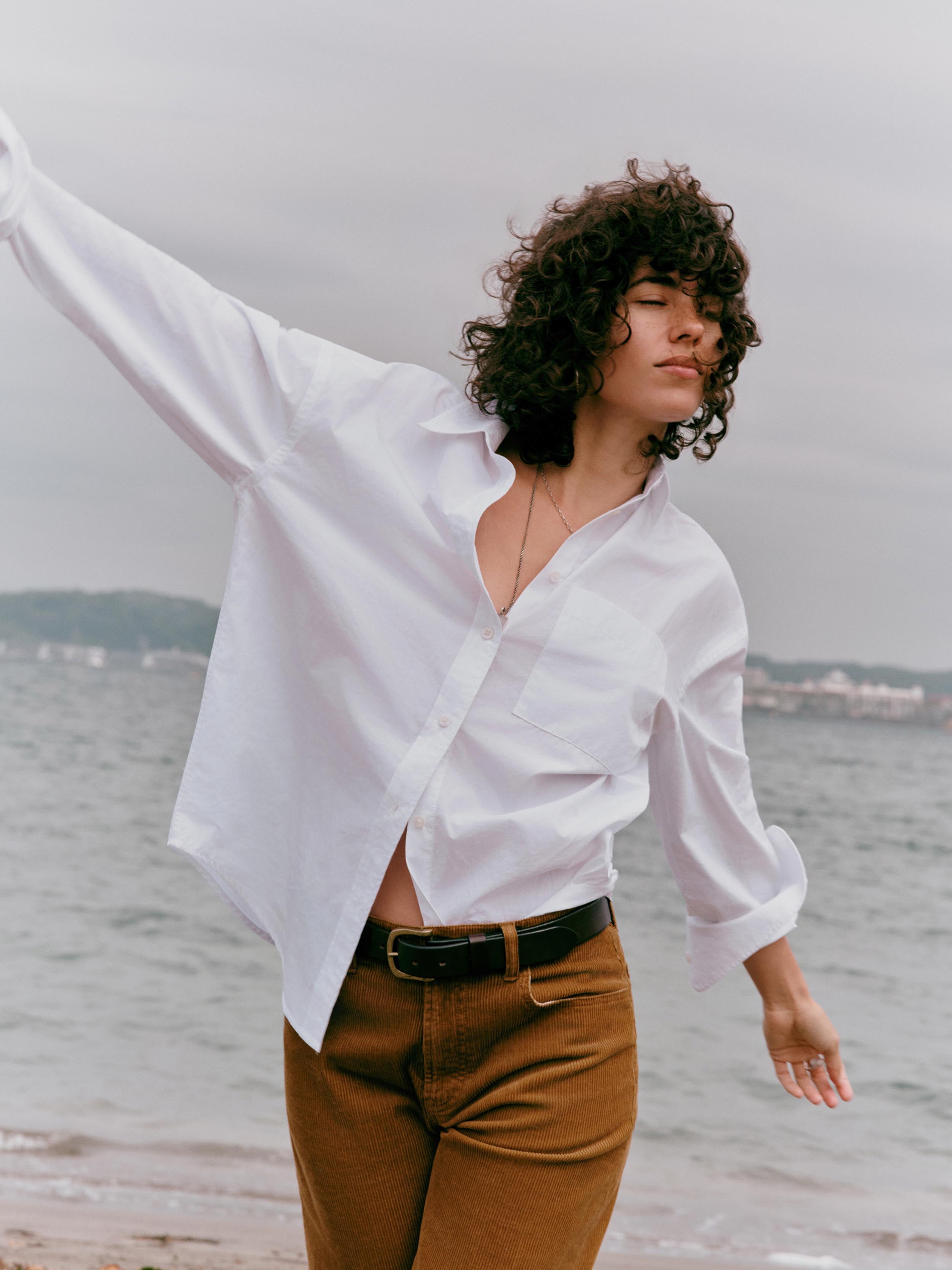A woman with curly dark hair stands on a beach wearing an oversized white button-down shirt and brown pants, with her arm raised against a cloudy sky and water in the background.