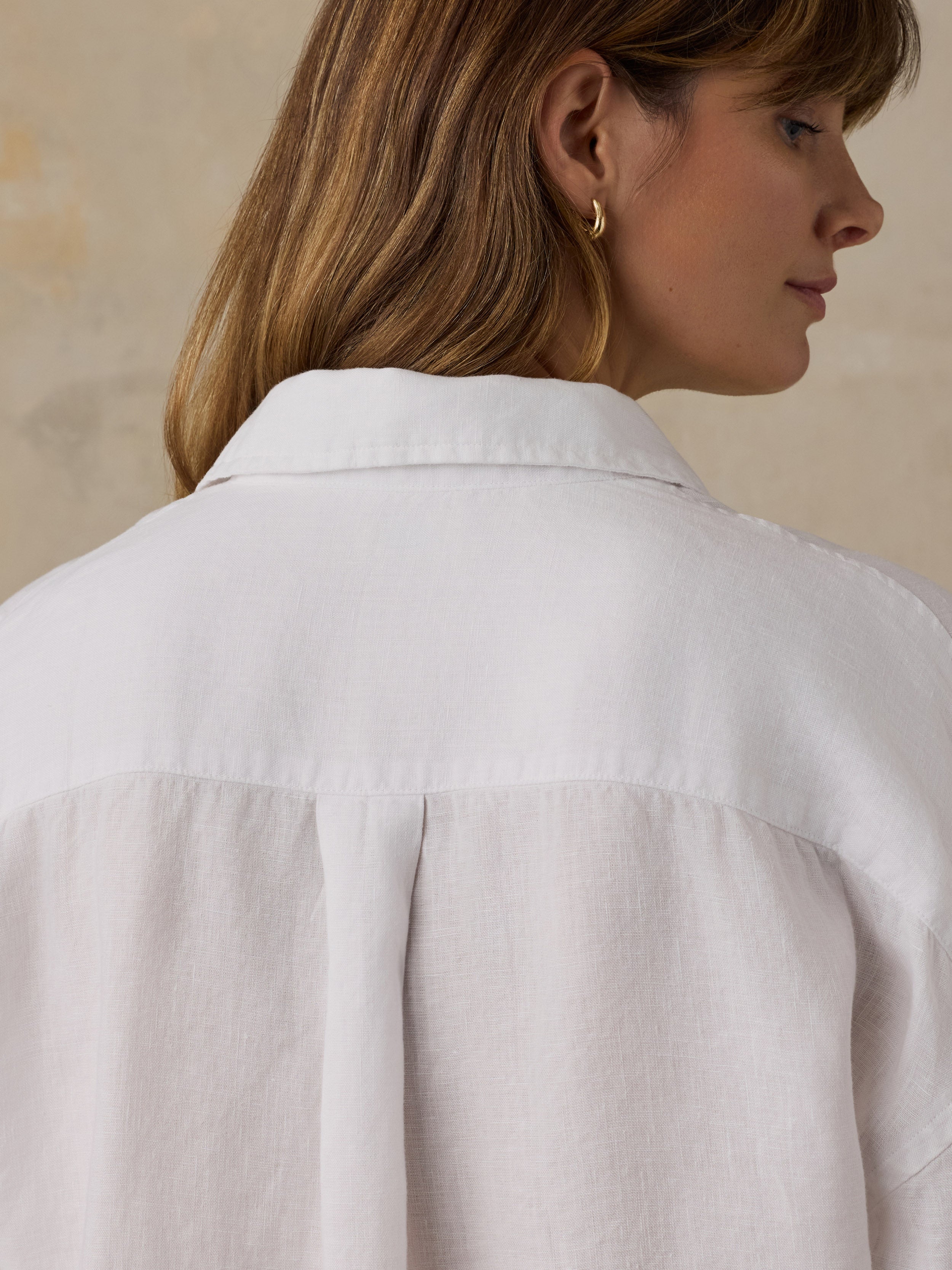 Close-up back view of a woman wearing a white button-up shirt, showing the collar, shoulder seams, and back fabric against a soft beige background.