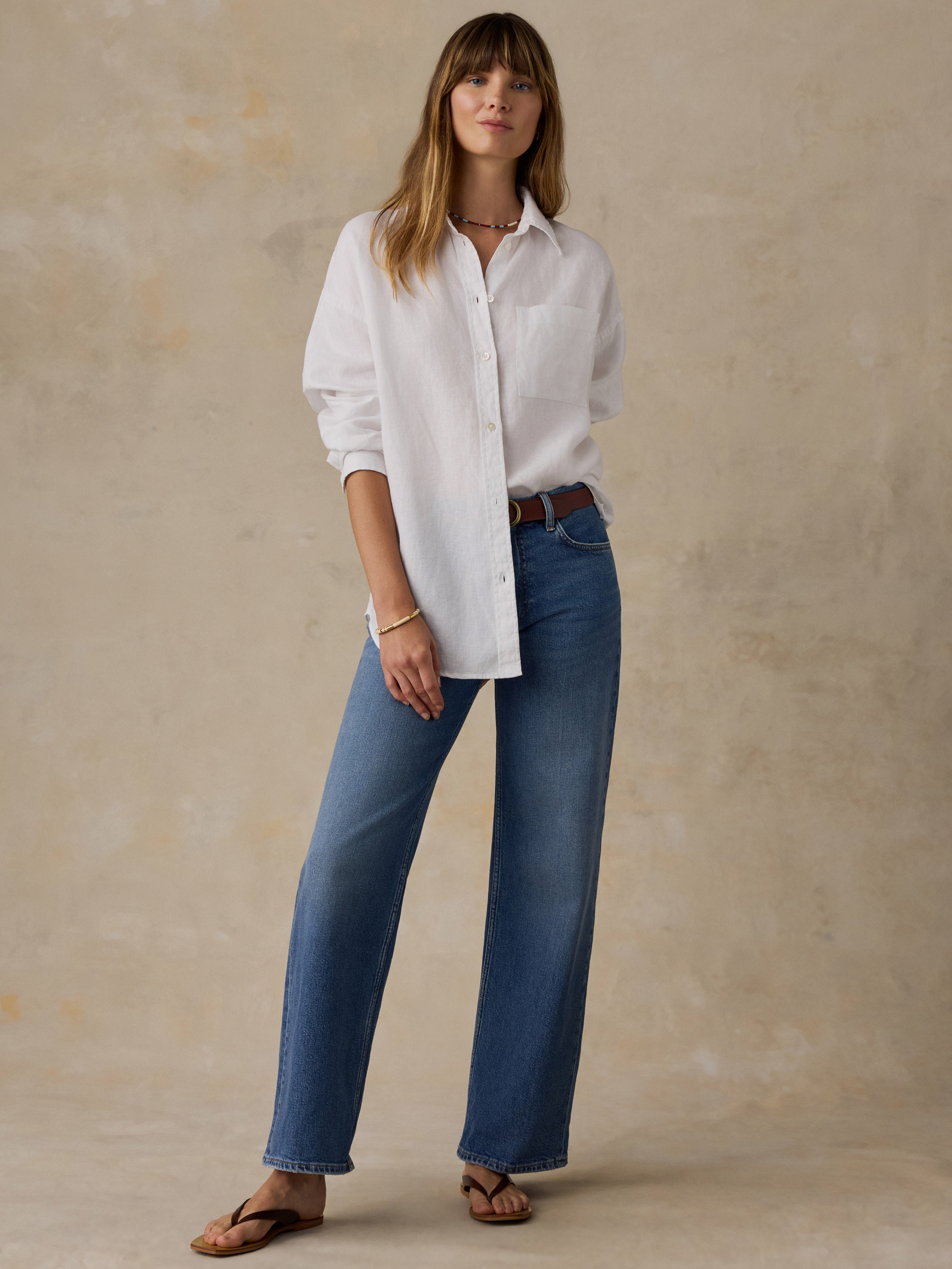 Woman wearing a white linen button down shirt with blue jeans against a beige background