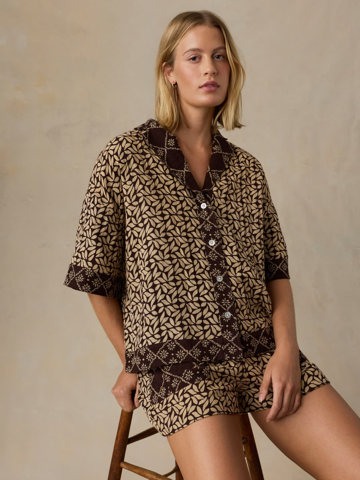Woman wearing a patterned outfit sitting on a stool against a neutral background, image 2 of 5