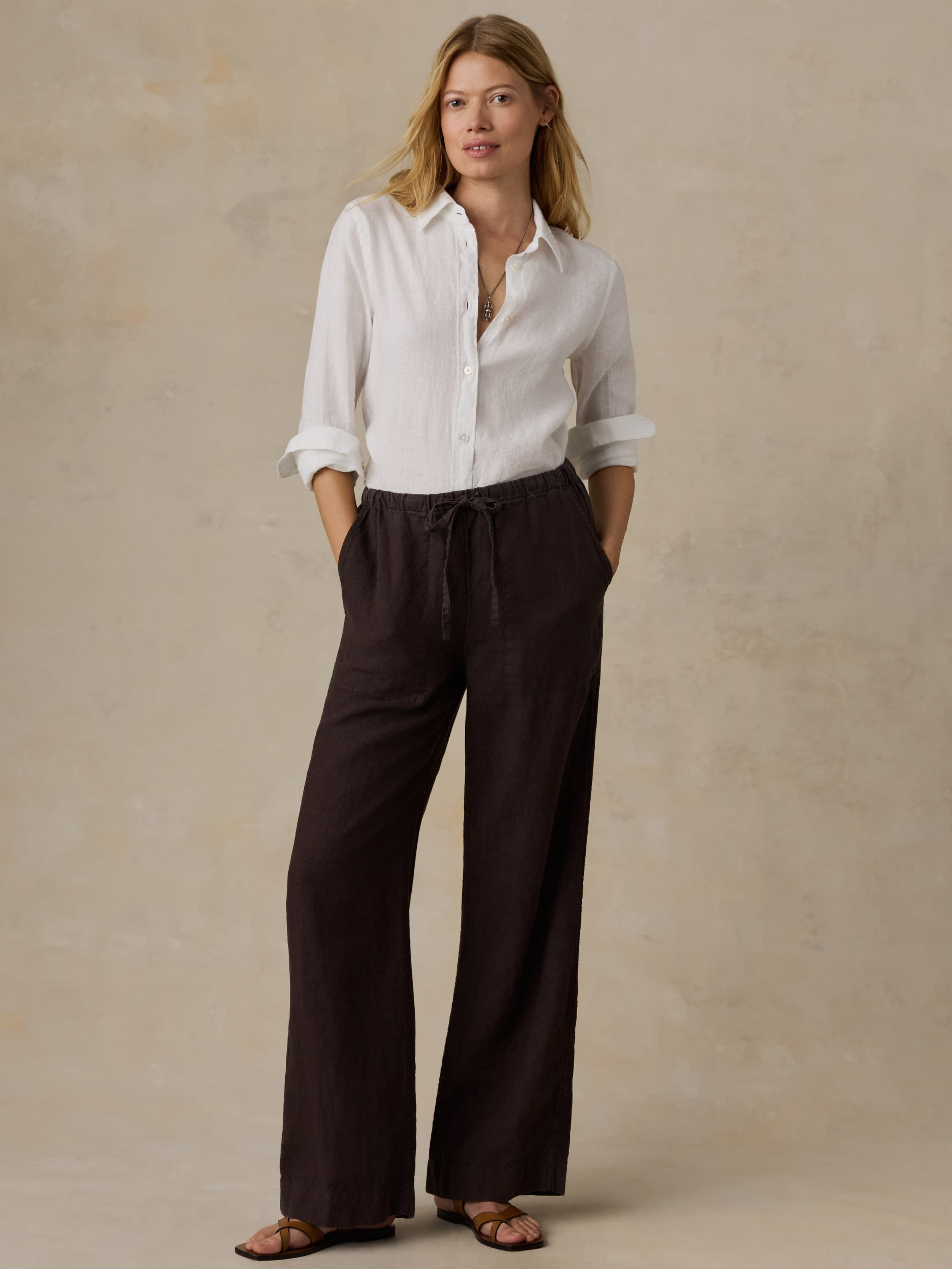 Woman wearing a white linen shirt and brown linen pants against a beige background