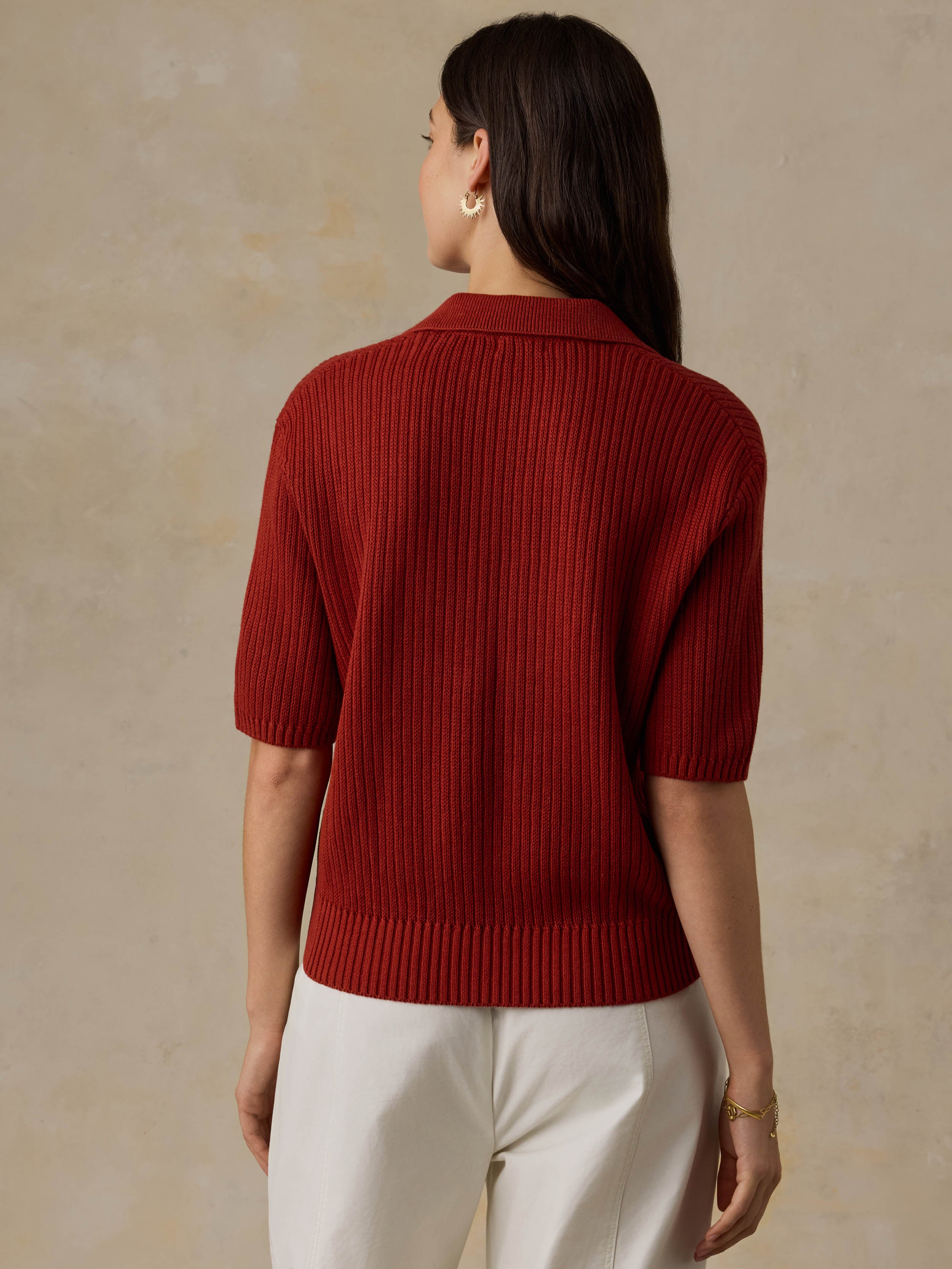 Woman wearing a red ribbed knit polo shirt and white pants against a beige background