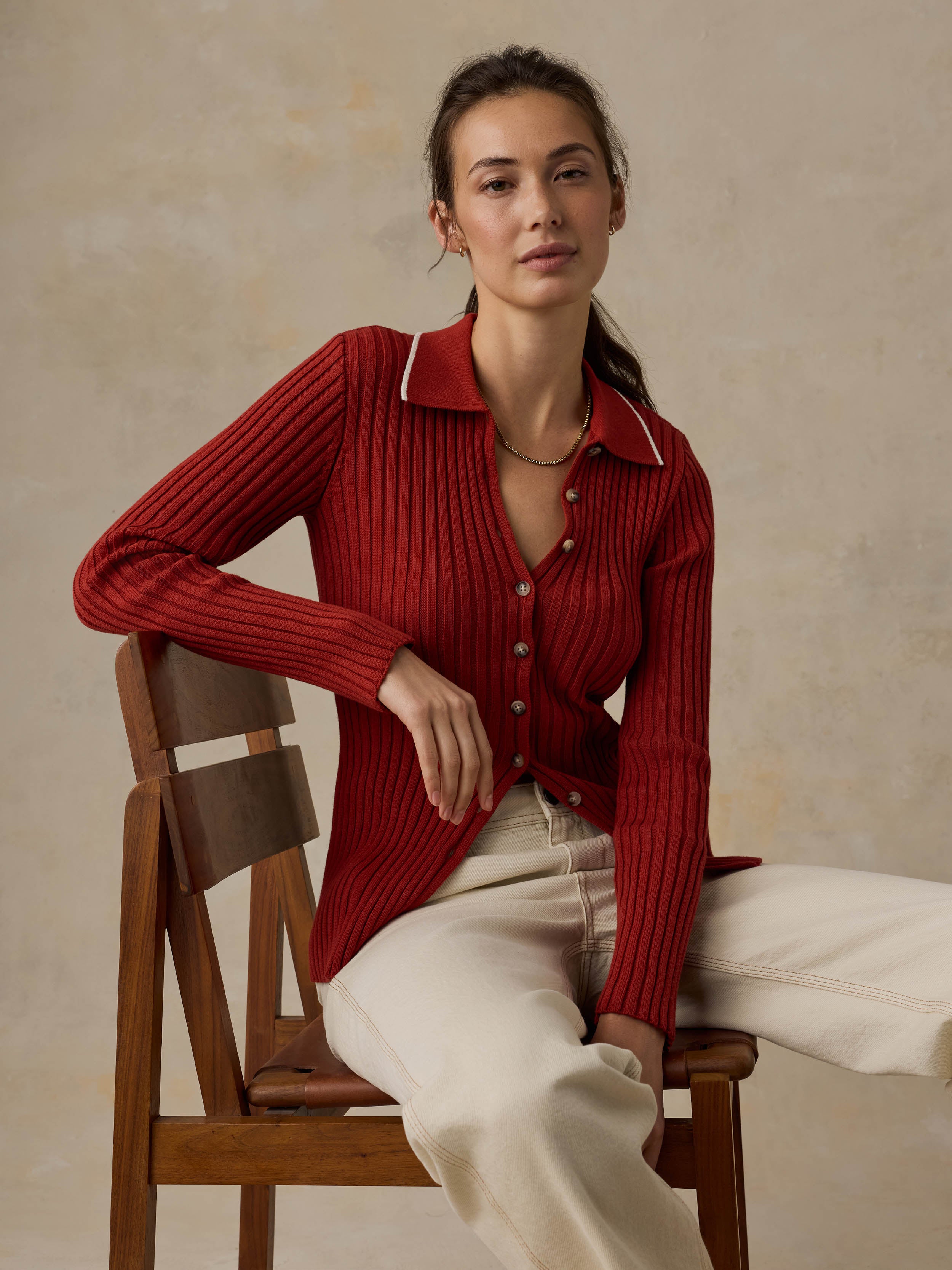 Woman sitting on a wooden chair wearing a red cardigan and white jeans against a beige background