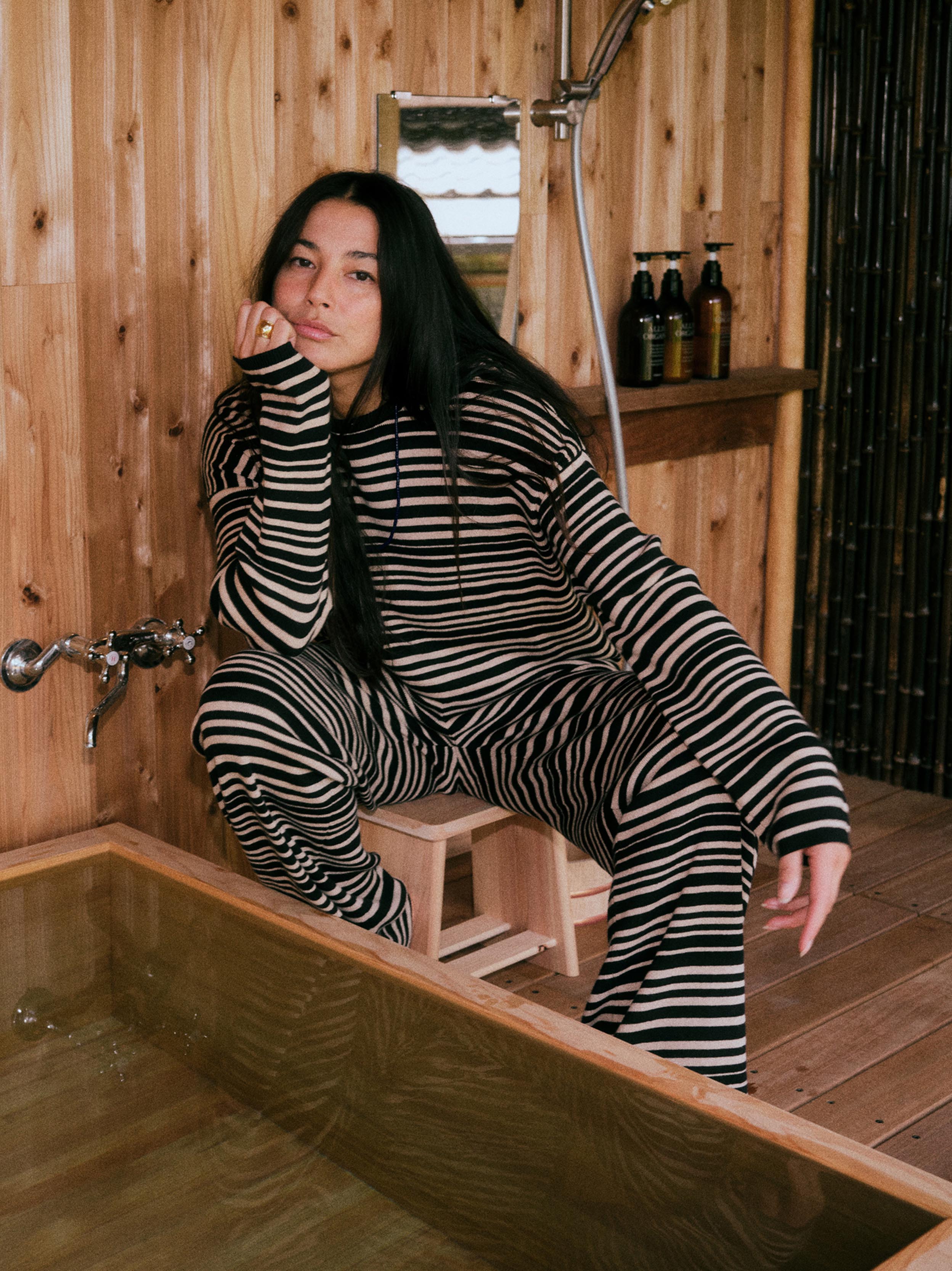 A woman with long black hair sits on a small wooden stool in a rustic wooden bathroom, wearing a matching black and beige striped lounge set. She is positioned next to a wooden bathtub, with a wooden wall, shower fixtures, and shampoo bottles in the background.